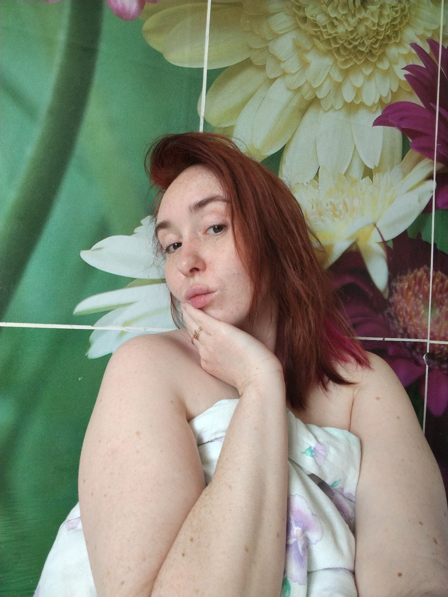 A person with red hair and visible freckles poses for a photograph indoors, wrapped in a floral-patterned blanket. The background features a floral design wallpaper or backdrop with green and white flowers. The person's face is angled slightly towards the camera, with their lips pursed in a kissing gesture. They are resting their chin on their hand, which has a ring on one of the fingers. The photograph appears to be taken in a well-lit area, showcasing the details of the person's features and the surrounding environment. No location of Krasnogorskoye, Russia is visible, so the location cannot be verified.