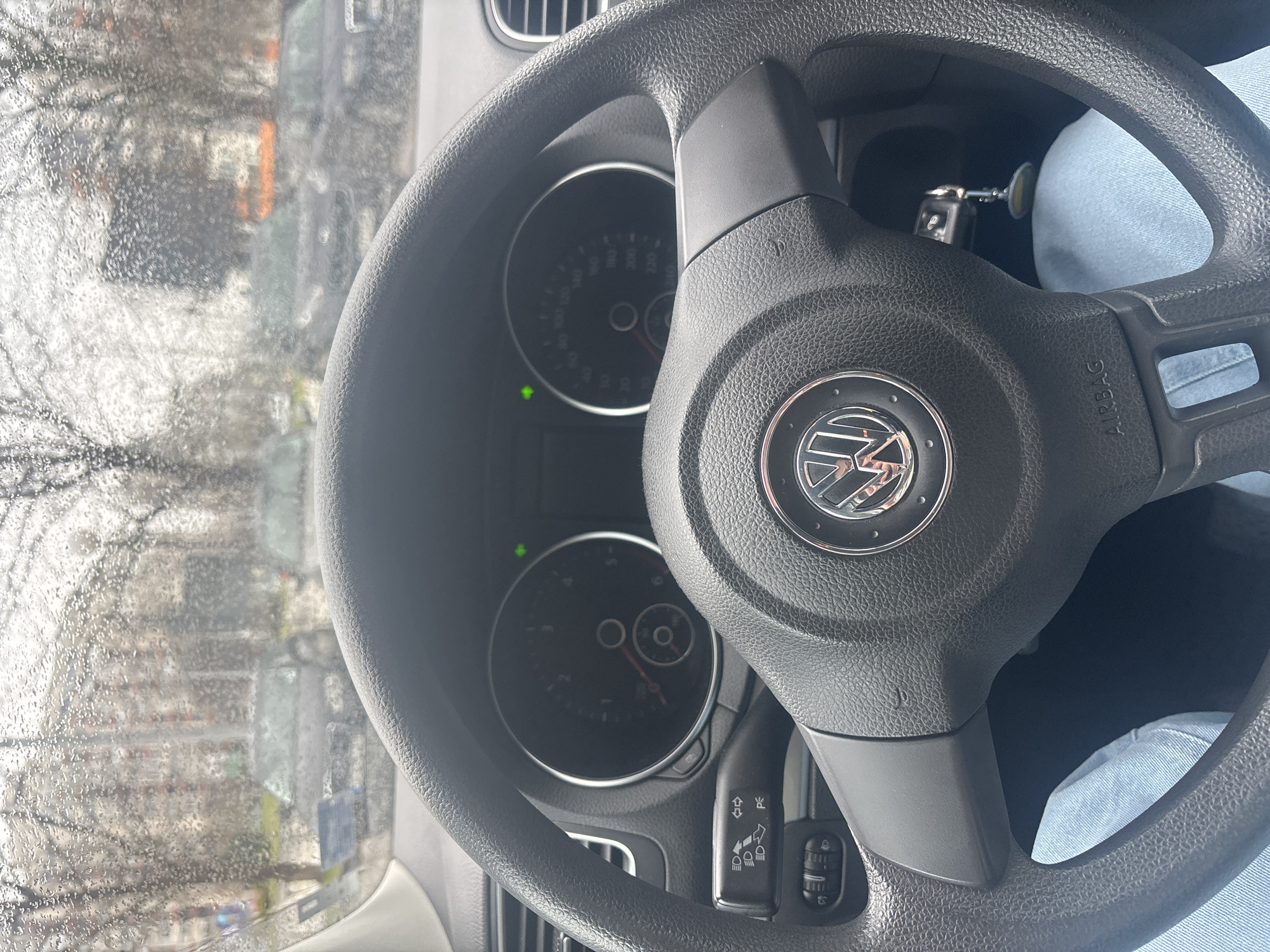 The image shows the interior of a vehicle, specifically the steering wheel of a Volkswagen car. The Volkswagen logo is prominently displayed in the center of the steering wheel. Behind the steering wheel, the car's dashboard and gauges are visible. Through the windshield, a blurred cityscape is visible. Based on the context provided, it can be inferred that the location is Plovdiv, Bulgaria. The weather seems wet, with raindrops visible on the windshield. A person wearing blue jeans is partially visible. A car key is visible.