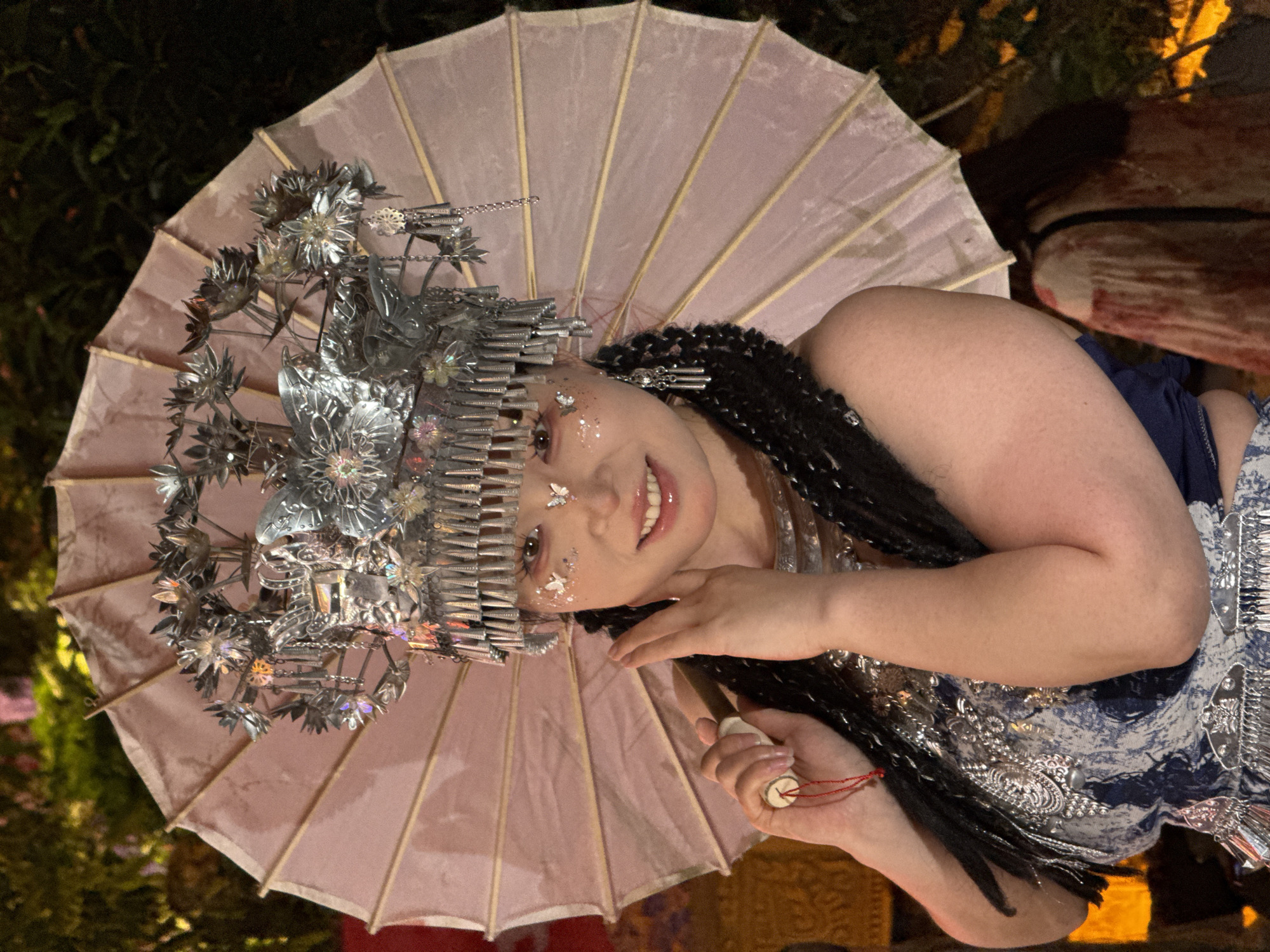 A woman is depicted, adorned with a silver headpiece featuring floral designs and what appear to be comb-like structures. She has embellishments on her face resembling small butterflies and is holding a light pink parasol. She is wearing a blue and white patterned garment. The location is dark with some vegetation. There is no confirmed connection to Yuzhong District, China.