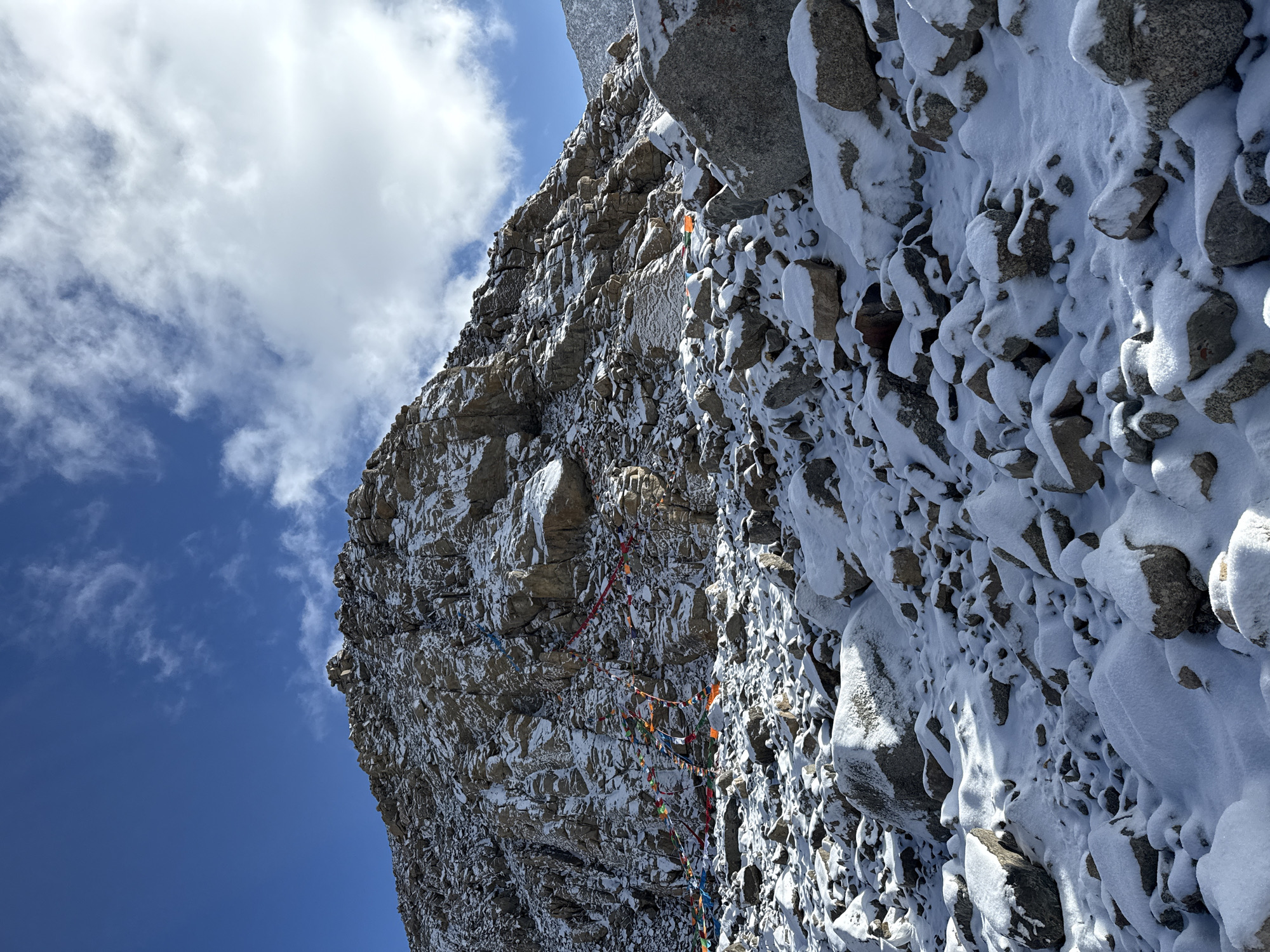 The image displays a majestic and rugged mountain face, heavily accented with patches of white snow and ice, set against a clear blue sky with scattered white clouds. The perspective is upward, emphasizing the sheer scale of the rock formations.
The primary subjects are the large, textured grey and brown rocks that form the steep cliff face. Snow clings to crevices, ledges, and uneven surfaces, creating a striking contrast with the darker rock. A notable object is the presence of numerous colorful Buddhist prayer flags (lungta) strung across the mountain. These flags, typically in blue, white, red, green, and yellow, are draped horizontally along the rocky contours, suggesting a path or a sacred site. They appear slightly faded, indicating exposure to the elements.
The setting is a high-altitude mountainous environment, consistent with regions known for spiritual practices involving prayer flags, potentially near the Parga Valley in China. There are no people visible, allowing the natural grandeur and spiritual markers to dominate the scene.
The image captures a moment of bright daylight, evidenced by the crisp blue sky and the way light and shadow define the rocky textures and snow. The weather appears clear and cold, suitable for snow to remain on the peaks. The overall impression is one of solemn beauty and spiritual significance. No visible text is present in the image.