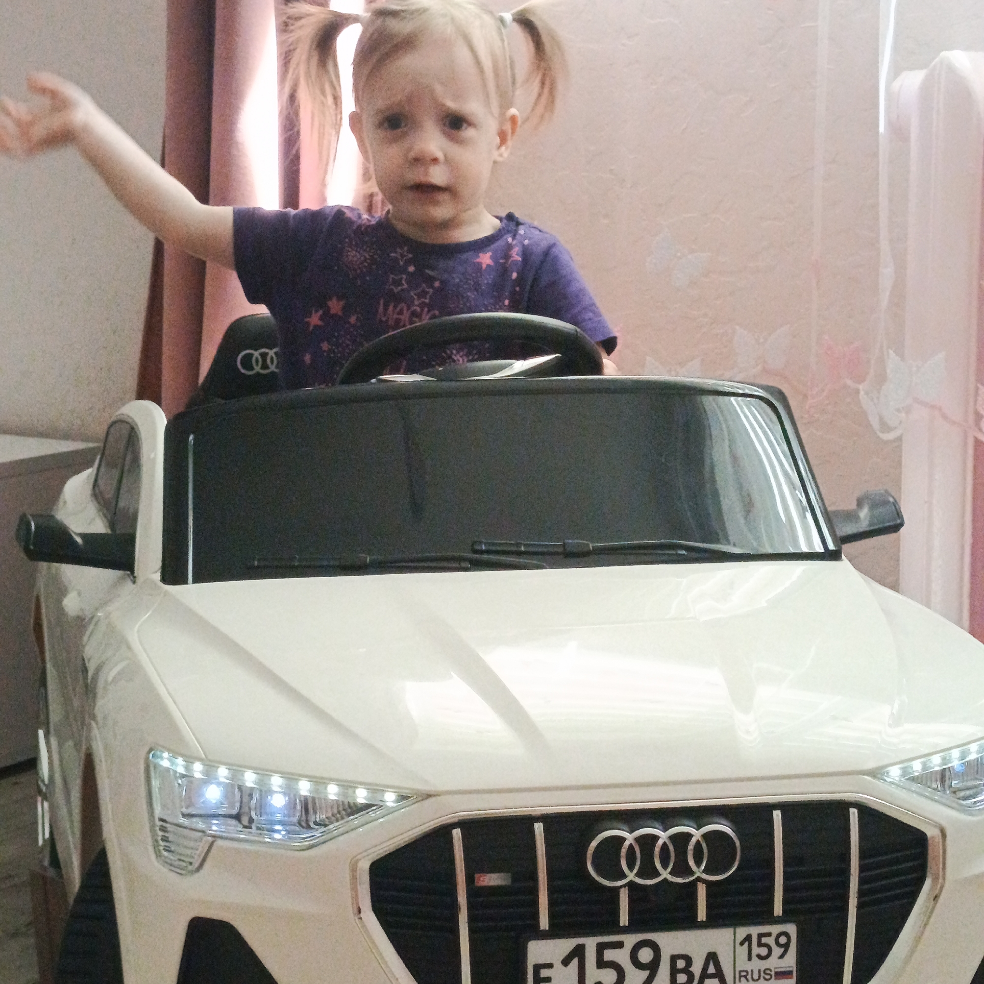A fair-skinned toddler with blonde hair in pigtails sits in a white Audi toy car. The toddler wears a purple t-shirt with stars and the word "MAGIC". The toy car features lit LED headlights, an Audi grille with four rings, and a Russian license plate. The background consists of pink curtains and wallpaper. The location is Марковский, Russia, based on the license plate.
