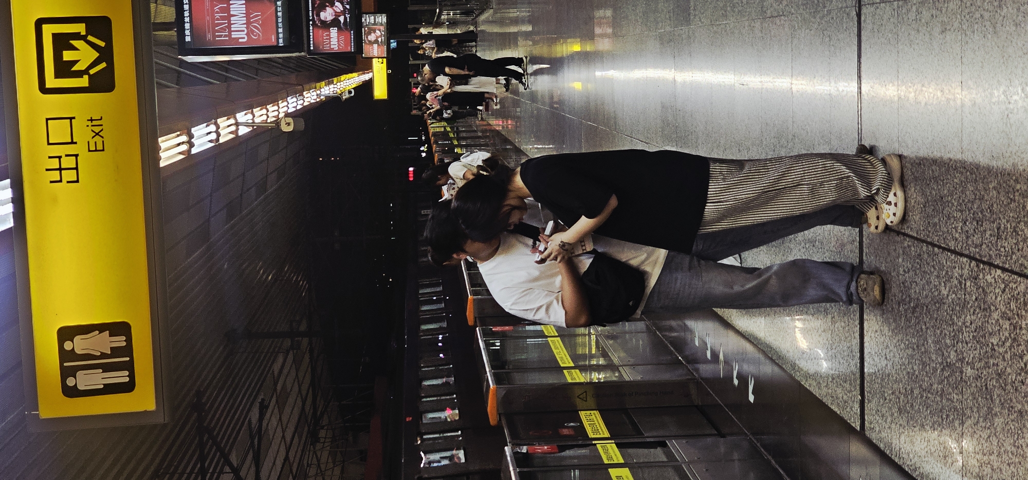The photograph depicts a couple embraced at a Chongqing High-tech Zone subway station in China. The woman is wearing a black shirt and vertically striped pants. The man is wearing a white shirt and gray pants. Both are looking at a cell phone, while standing adjacent to the subway platform glass enclosure that displays "Caution of Punching Habit". Signs indicate "Exit" and "Bathroom" with symbols. Other people stand along the platform. Advertisements are visible along the top of the opposite wall. The lighting is dim.