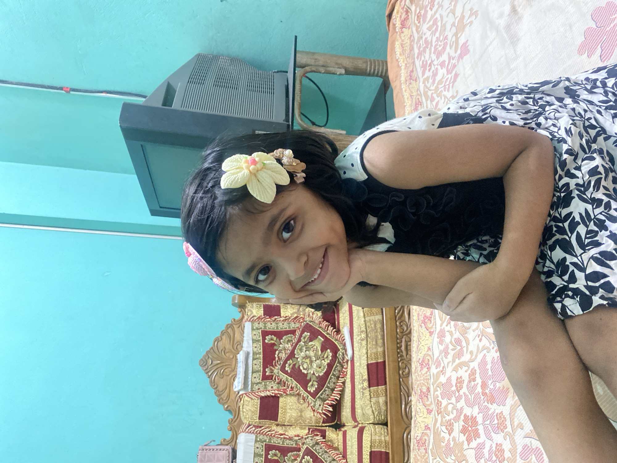 A young South Asian girl is sitting on a bed in a room. She is wearing a sleeveless dress with a black top and a white skirt with black floral print. She has a flower hair clip in her hair. She is smiling and resting her chin on her hand. The background shows a wooden sofa with red and gold cushions and a television set. The walls are painted a light blue color. The lighting is artificial and appears to be from an overhead source. The room is likely a living room or bedroom in a residential dwelling.