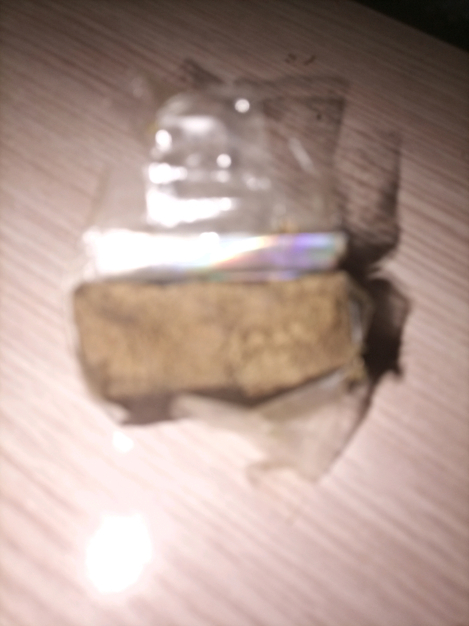 The image shows a blurry, close-up view of a rectangular object, possibly a brick or a block, wrapped in clear plastic. The object is placed on a surface that appears to be a wooden table with a wood grain pattern. The lighting in the image is uneven, with bright highlights and shadows, suggesting an indoor setting. There are no people or discernible landmarks that would indicate a specific location in Tarragona, Spain, or any other city. The overall impression is that of a simple, unadorned scene, focused on the object itself.
