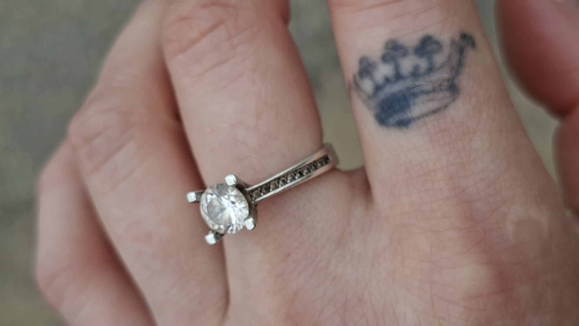 The media file shows a close-up view of a person's hand, with a ring on one of the fingers. The ring features a large, clear stone set in a silver band, with smaller dark stones lining the sides of the band. On the finger adjacent to the one wearing the ring, there is a faded tattoo of a crown. The person's skin appears pale, with visible lines and some lighter patches, possibly indicating scarring or a healed injury. The background is blurred, suggesting a shallow depth of field, and appears to be a neutral, muted color, possibly a wall or fabric.
The image seems to capture a moment of personal expression or adornment, focusing on the detail of the ring and the tattoo. There is no discernible time of day or weather information. The setting is not clearly defined beyond it being an indoor or close-up environment. No text is visible in the image. The overall tone of the image is neutral and observational, focusing on the visual elements of the hand, the jewelry, and the tattoo. The location context provided, Забалоцкі сельскі Савет, Belarus, does not offer direct visual cues within the image itself.