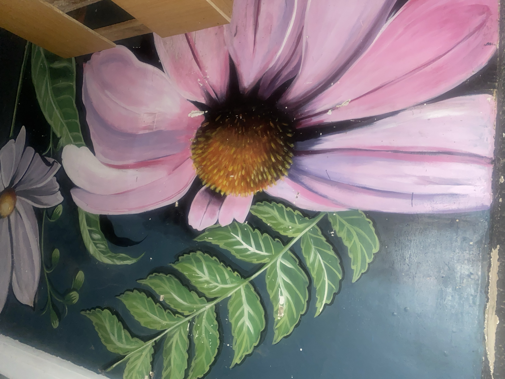 The image is a close-up of a mural depicting large flowers and green leaves against a dark teal background. A prominent pink coneflower with a textured brown and orange center is in the foreground, its petals spanning across the frame. To the right, a portion of a light purple daisy-like flower is visible. Green fern-like leaves are positioned above and behind the flowers. The mural appears to be painted on a rough, textured wall, with some visible damage and peeling paint along the left edge. In the lower right corner, two unvarnished wooden planks are placed horizontally, partially obscuring the mural. The setting is an interior space, potentially a wall in a building within the city of Howayi, Nigeria. There are no people or discernible actions taking place in the image.