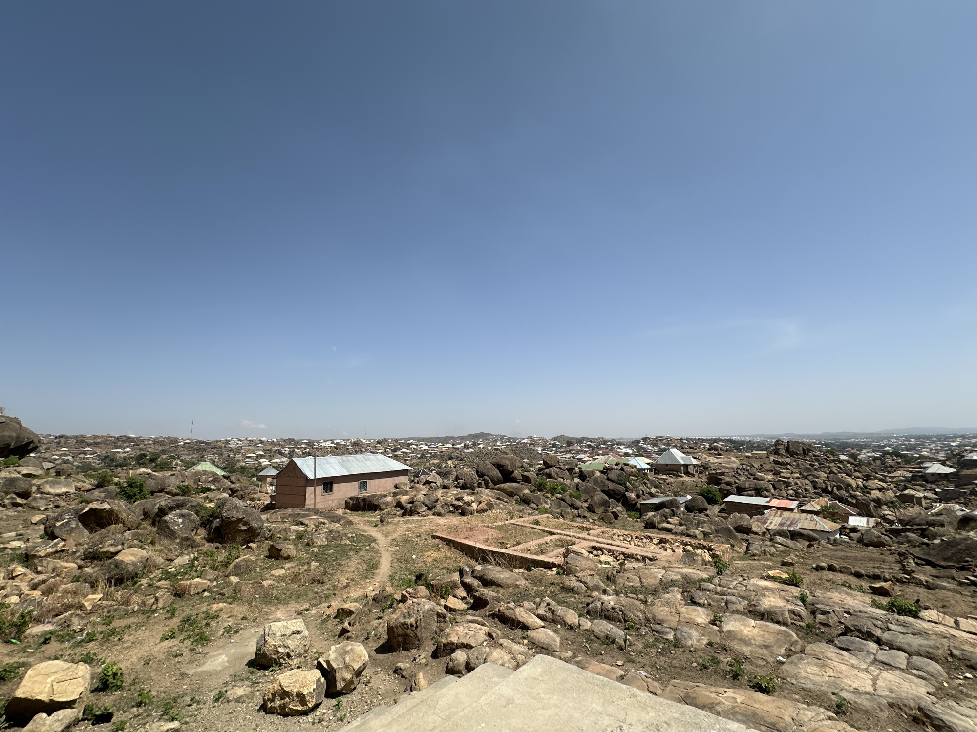 The image depicts a rocky landscape with scattered buildings, likely in the city of Jos, Nigeria. Numerous large boulders are present, interspersed with dry vegetation and paths. A few single-story buildings with corrugated metal roofs stand among the rocks. The sky is clear and blue. In the distance, a dense collection of buildings suggests a populated area. In the foreground, a concrete structure is visible, possibly part of a staircase or platform.