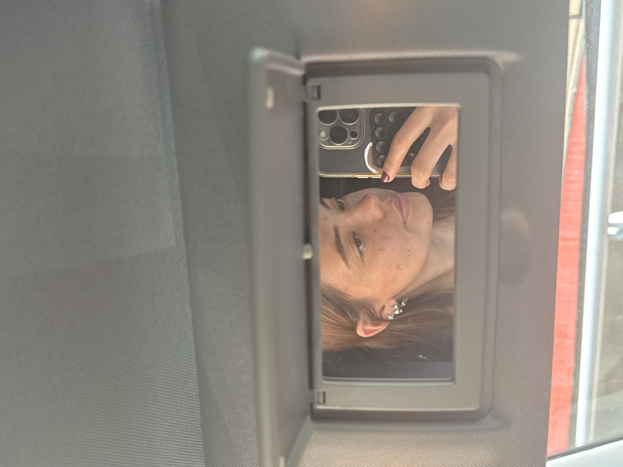 A selfie is taken in the mirror of a car's sun visor, which is flipped down. The selfie shows a woman's face, partially obscured by a phone, with her hand visible in the reflection, holding the phone. The woman has freckles and multiple earrings in one ear. The background appears to be the interior of a car, with a view out of the window showing a red structure partially visible, potentially in Puebla, Mexico.