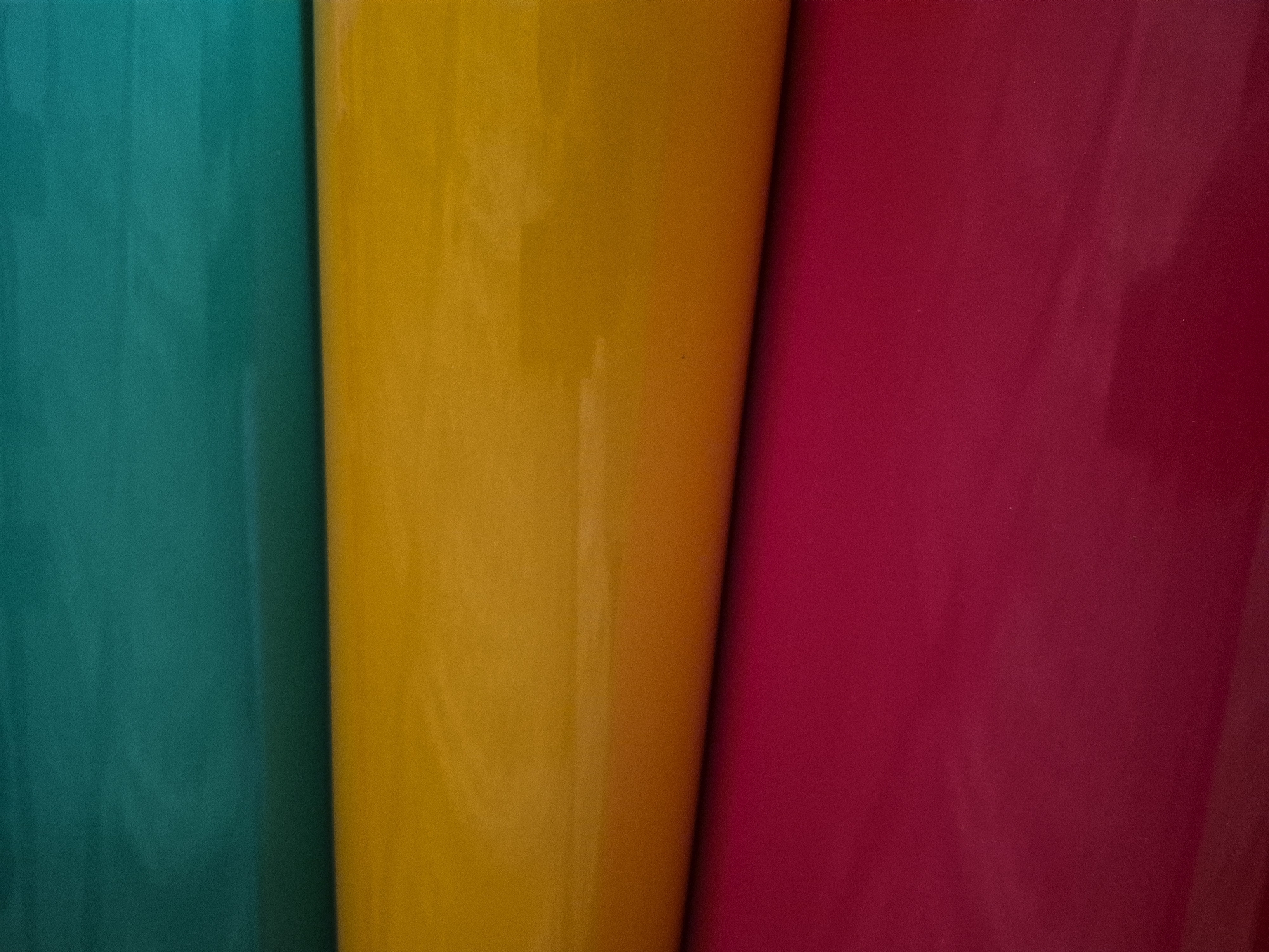 The image shows a close-up, horizontal view of three rolled-up pieces of fabric, stacked on top of each other. The top fabric is a deep teal color, the middle fabric is a bright yellow-orange, and the bottom fabric is a vibrant magenta. The rolls are positioned diagonally from the upper left to the lower right of the frame. The lighting creates subtle shadows and highlights, suggesting a slightly textured surface on the fabrics. The background is dark and out of focus. This image does not depict any specific location in Masów, Poland.