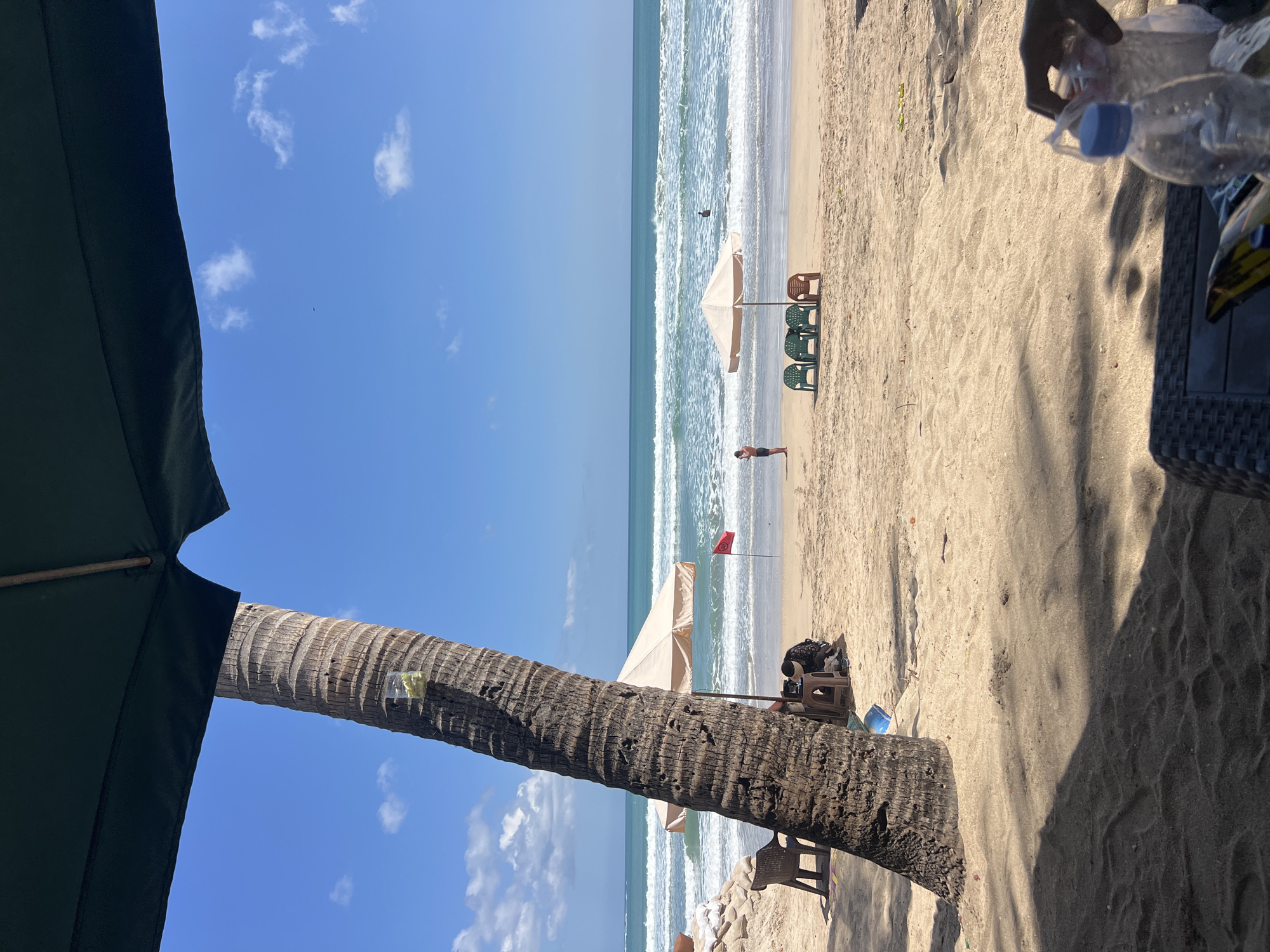 The image depicts a sunny day at Kuta Beach in Indonesia. In the foreground, the shadow of an umbrella stretches across the sand, partially obscuring a dark-colored table with a plastic water bottle and other items. A person's hand is visible reaching for the items. The middle ground features a large palm tree trunk on the left, with a clear plastic cup containing some green items hanging from it. To the right of the palm tree, a beach umbrella provides shade over a few chairs. Further out, a person stands near the water, and another individual is swimming or wading in the ocean. The ocean itself shows moderate waves breaking on the shore under a clear blue sky dotted with a few white clouds. A red flag is planted in the sand near the water's edge, suggesting potential safety warnings. The beach is sandy, with some ripples and footprints visible.