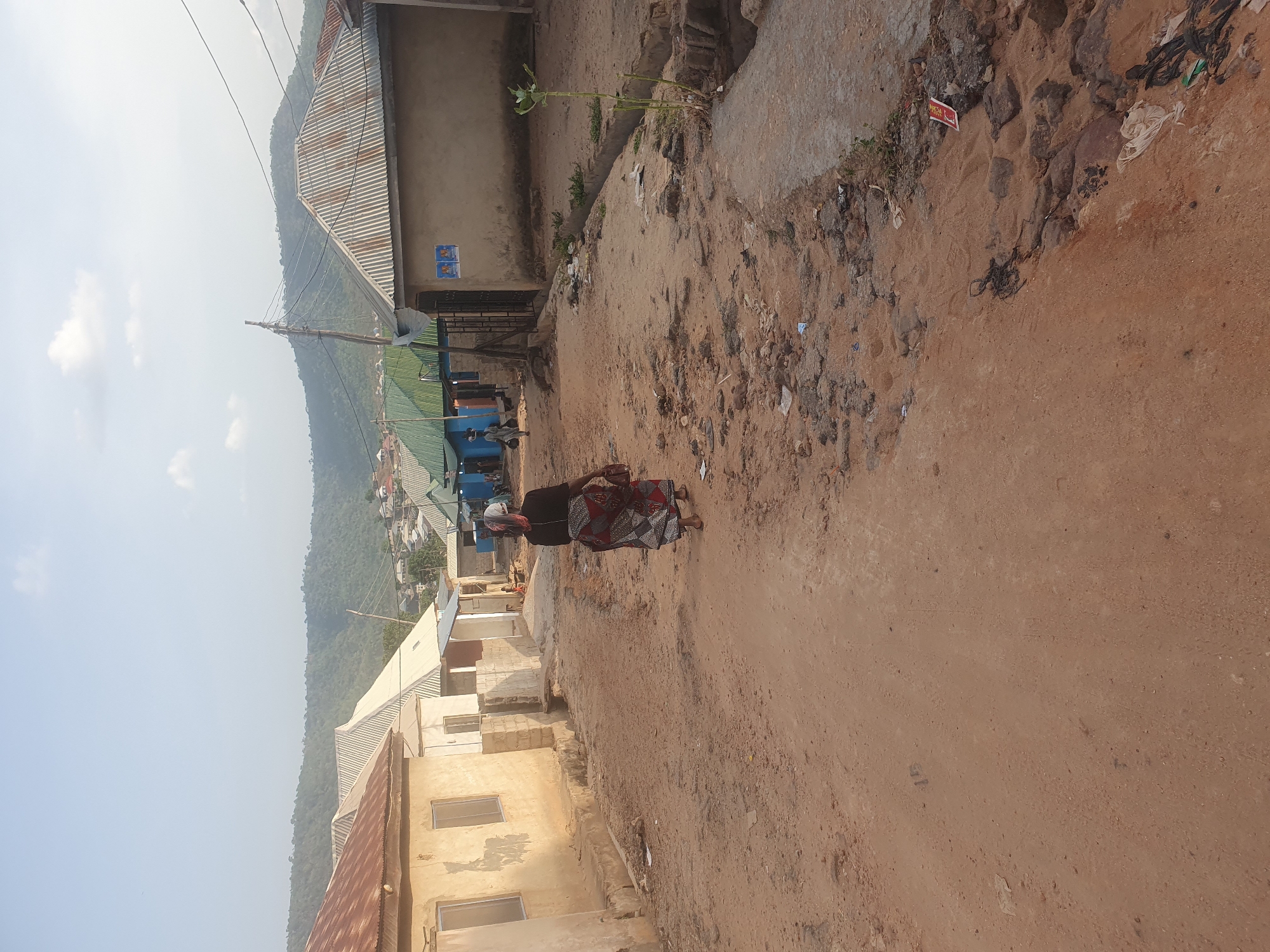 The image depicts a dirt road winding through what appears to be a residential area in Lokoja, Nigeria. The houses lining the road are a mix of single-story structures, some with corrugated metal roofs and others with more finished facades. A woman is walking along the road, wearing a dark top and a patterned skirt. The landscape in the background includes a hill covered in vegetation. The overall environment suggests a lower-income or developing area within the city.
