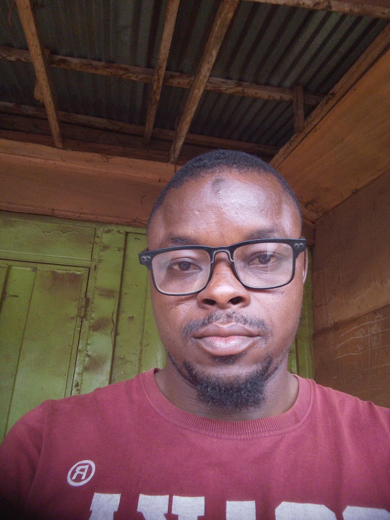 A man is posing for a selfie in an indoor space. He is an African man, wearing black framed glasses, a maroon t-shirt with a white logo on the left, and a small beard. The background includes a green door, a brown wall, and a corrugated metal roof supported by wooden beams. The specific location in Tamale, Ghana cannot be determined from the image.