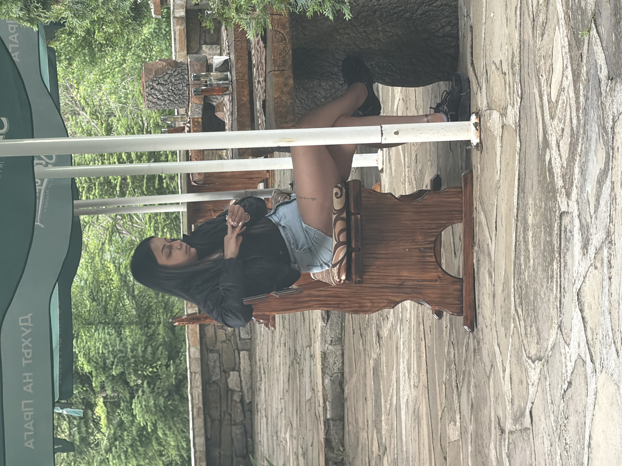 An outdoor scene in Sushina, Bulgaria, shows a woman sitting on a rustic wooden bench or chair under the shade of a large green umbrella. The woman, with long dark hair, is dressed in a dark long-sleeved top and light denim shorts, with black sneakers on her feet. Her legs are crossed, and she appears relaxed, with her hands clasped in front of her. Rings are visible on her fingers, and a small, dark tattoo can be seen on her upper left thigh.
The setting suggests a cafe or restaurant patio. Behind the woman, a sturdy wooden table or structure, possibly made from a tree trunk, holds several glass bottles, likely containing spirits or condiments. The background is lush with green foliage, indicating a natural, perhaps garden-like, environment. The ground and surrounding structures consist of irregular stone paving and textured stone walls, contributing to a natural, rustic aesthetic.
Visible on the green umbrella, in Cyrillic script, is the text "ПАЛАЦ НА ДУХА," which translates to "Palace of the Spirit." The time of day appears to be daytime, with ample natural light suggesting clear weather. The overall mood is calm and serene.