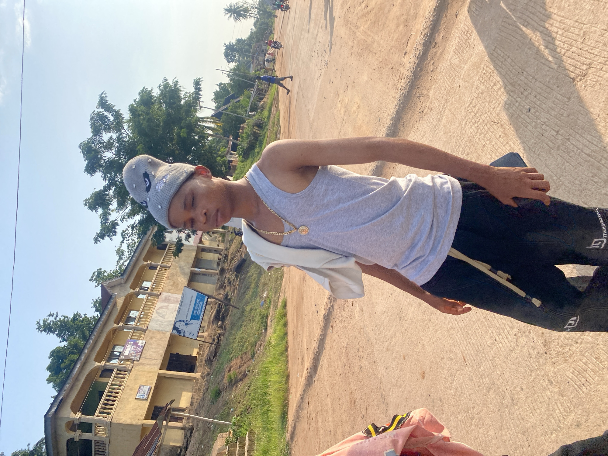 The image depicts a rural or semi-urban street scene in Nguzu Edda, Nigeria, under bright daylight conditions. In the foreground, slightly left of center, stands a young male. He has light skin, dark hair (mostly covered), and is wearing a light gray knit beanie adorned with a stylized, angry-looking character design featuring black eyes, a white eyebrow, and studded accents. He wears a light gray tank top and black trousers with a light-colored drawstring or belt hanging from the waist. A prominent gold chain with a circular gold pendant rests on his chest. His eyes are closed or heavily squinted, and his head is slightly tilted. His left hand, with a dark-colored object resembling a phone or wallet, rests against his leg. A white garment is draped over his right shoulder.
In the midground and background, an unpaved, light-brown dirt road extends into the distance, showing some faint tire track patterns. To the left, a grassy hillside with steps leads up to a two-story building. This building features yellow-painted walls, an elaborate balcony with balustrades on the upper level, and multiple windows. A sign partially obscured but reading "DUNAMIS" is visible on the upper floor balcony. Another large blue and white sign, possibly an advertisement or political poster, is positioned on the ground in front of the building. Further down the road, several individuals, mostly adult males, are visible. One man in a blue shirt walks towards the left, while others on motorcycles are further in the distance. Palm trees and other green foliage are scattered throughout the background under a clear, light blue sky with minimal cloud cover. The overall impression is a sunny, warm day in a developing community.