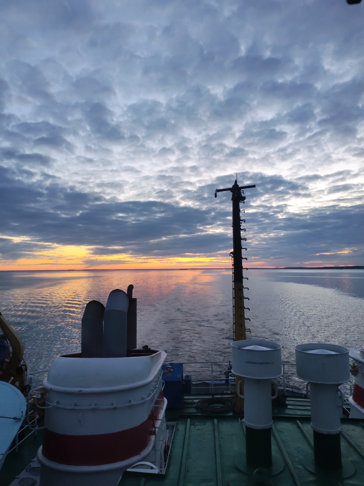 The image captures a serene view from the stern of a ship sailing on a calm body of water, likely at sunrise or sunset, given the warm light on the horizon. The sky is mostly covered with dense, textured clouds, creating a dramatic backdrop. Towards the distant horizon, a vibrant band of orange and yellow light stretches across the sky, reflecting beautifully on the water's surface.
In the foreground and midground, various parts of the ship are visible. On the left, a large structure with white and red banding, possibly part of a funnel or superstructure, can be seen, with two grey exhaust stacks rising behind it. On the right, a tall, dark mast, equipped with numerous rungs or steps and horizontal crossbars, stands prominently against the colorful sky. A light fixture or sensor is attached near the top of the mast. The ship's movement is indicated by the distinct wake trailing behind it in the water.
On the green-colored deck in the lower right, two white, cylindrical vent-like structures are present. One of these structures clearly displays the Russian text "АККУМУЛЯТОРНАЯ", which translates to "Battery Room" or "Accumulator." The scene is devoid of people, emphasizing the vastness of the water and sky. The tranquil setting and soft, low light evoke a peaceful atmosphere near Gorushka, Russia.