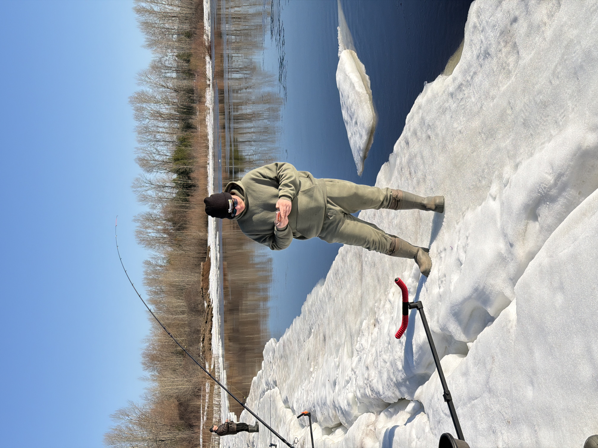 The photograph depicts a man standing on a snowy bank beside a river. He is wearing a dark beanie, a light green hooded sweatshirt, light green pants, and rubber boots. He has a mask pulled down around his chin. He is holding something small in his hands and looking down.
A fishing rod is set up with a rod holder on the snow bank. More fishing rods are set up further down the bank with another person standing near them. The river behind the man is partially covered with ice floes. On the opposite bank, there are bare trees. The sky is blue and clear.
The location is Chaikovsky, Russia.