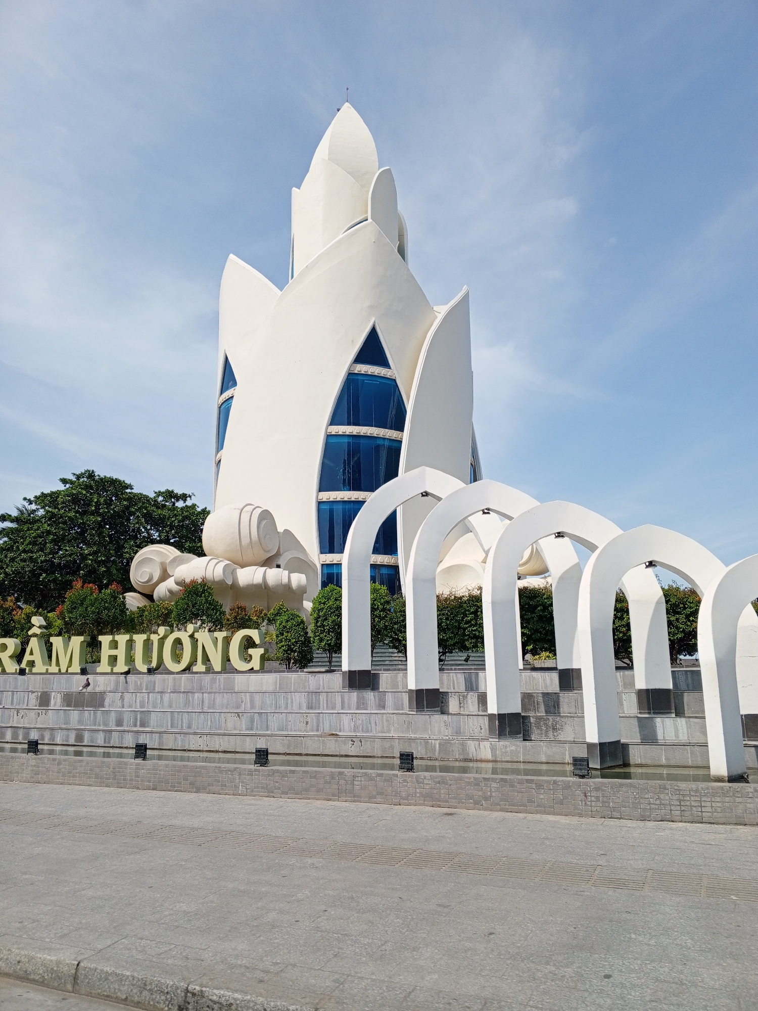 The image shows the Nha Trang Center for Cultural Tourism, also known as the Tram Huong Tower, located in the city of Nha Trang, Vietnam. The tower is a modern architectural structure with a distinctive lotus-like design, featuring white petal-shaped elements and large blue glass windows. In the foreground, there is a large sign with yellow lettering that reads "RÀM HƯƠNG". This sign is placed in front of a tiered structure with stone steps and a water feature. A row of white arched structures, resembling a colonnade, stands in front of the tower. Lush green trees and shrubs are visible behind the sign and the arched structures, creating a vibrant contrast with the white architecture. The sky is a clear blue with a few wispy clouds, indicating a sunny day. The scene is captured from a slightly low angle, emphasizing the grandeur of the tower and the surrounding features.