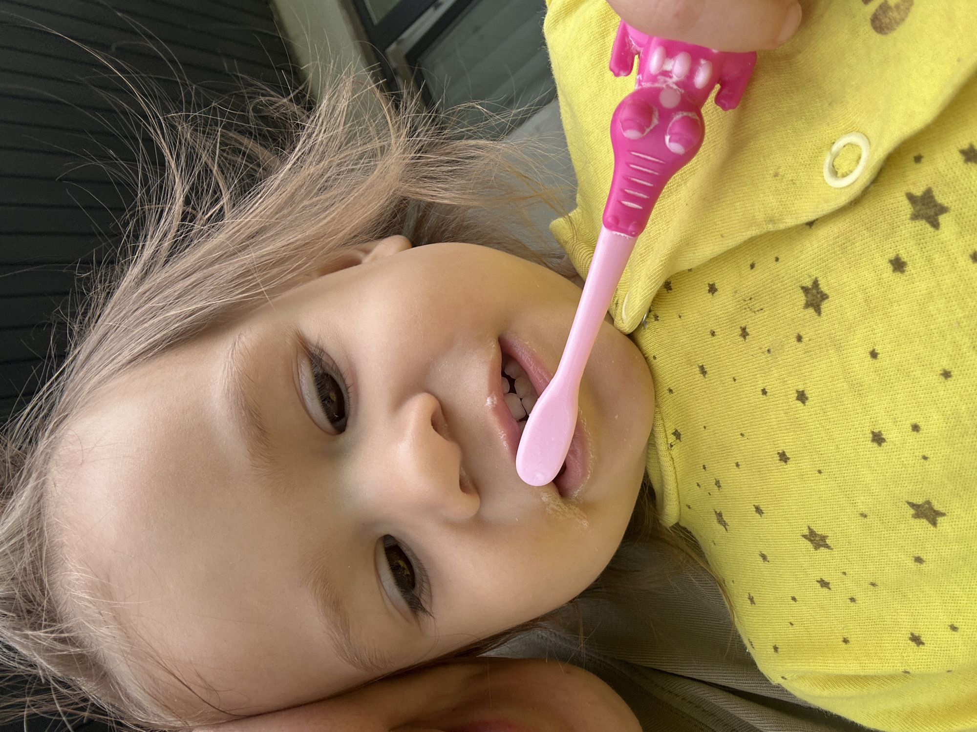 A toddler with blond hair is lying down and holding a pink elephant-shaped toothbrush in her mouth. She is wearing a yellow button-down shirt with brown star patterns. The background is blurred. The location is not discernible from the image.