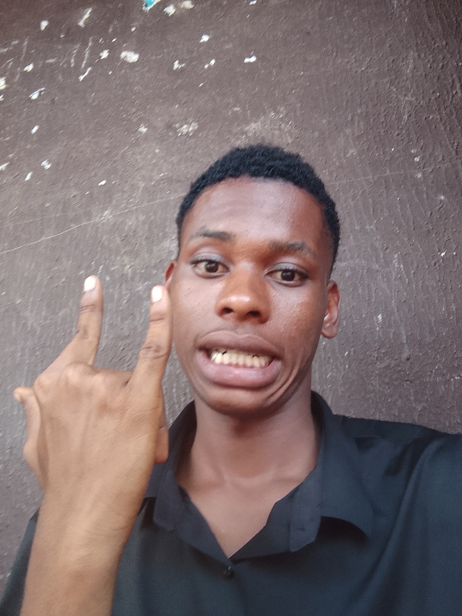 A young Black man is posing for a selfie, making the "rock on" hand gesture. He is wearing a dark-colored collared shirt. The background is a wall with a mottled brown texture. The picture was taken in Nkpor, Nigeria.