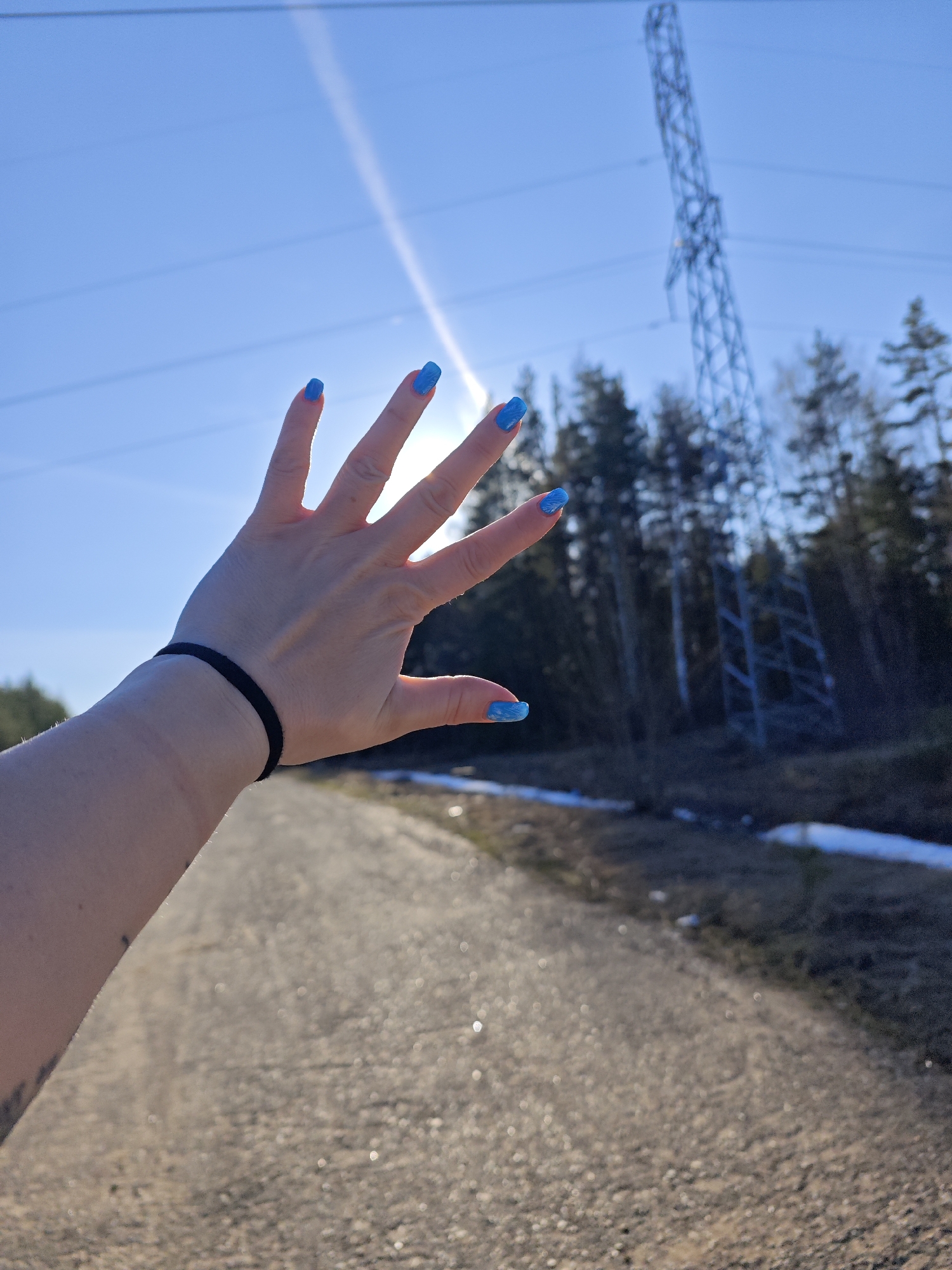 The image presents an outdoor scene in Chaikovsky, Russia, captured under bright daylight with a clear blue sky. In the foreground, a person's right hand and forearm are prominently featured, with the hand held up towards the sky. The fingernails are adorned with a vibrant, bright blue nail polish. A black hair tie is worn around the wrist.
The hand appears to be either shielding from the strong sunlight, which emanates from behind it, or performing a gesture. Strong sun rays are visible, creating a bright glare in the upper right portion of the image.
The background reveals a natural and infrastructural landscape. A dirt or gravel path extends into the distance on the right, flanked by patches of melting snow, indicative of late winter or early spring. Beyond the path, a dense forest of evergreen trees is visible. A tall, metal electricity pylon or transmission tower stands prominently among the trees in the mid-ground, with power lines stretching across the clear blue sky. No specific activity beyond being outdoors and holding up the hand is discernible. There is no visible text in the image.