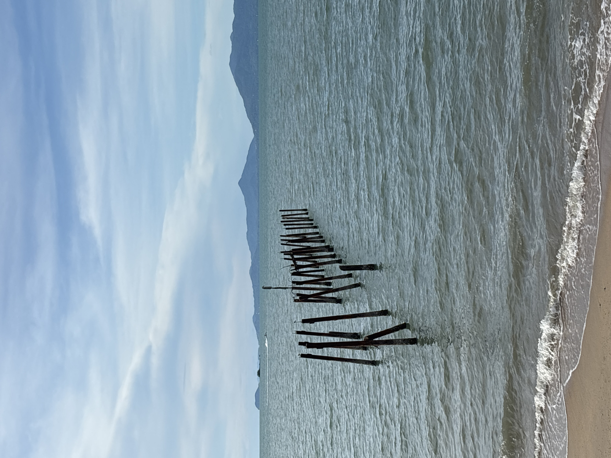 The image captures a dilapidated wooden pier extending into the sea at Baan Bo Phut, Thailand. The pier is in a state of disrepair, with many planks missing and the supporting structures broken. The water is calm, with gentle waves lapping against the shore. In the background, the silhouette of mountains is visible. The sky is partially cloudy. The scene suggests a quiet, undeveloped beach area.