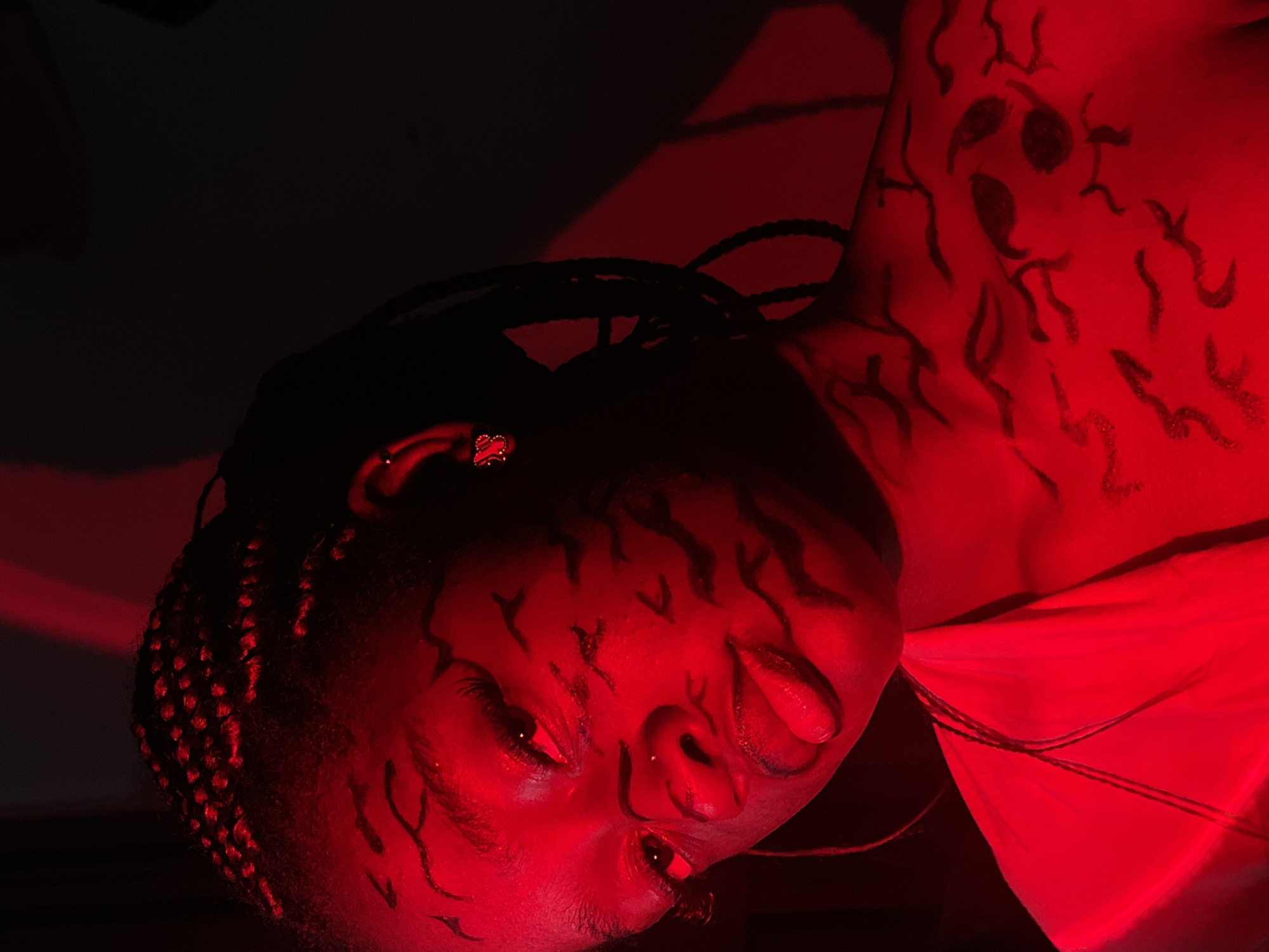 A close-up portrait of a person with dark skin, possibly female, with braided hair. The person is lit with a strong red light, causing most of the scene to appear red. There are dark markings on the person's face and chest resembling stylized tribal or abstract patterns. The person is wearing an earring with a red design. The background is mostly dark and out of focus. The location is unknown and there are no clues to locate the city of Ado, Nigeria.