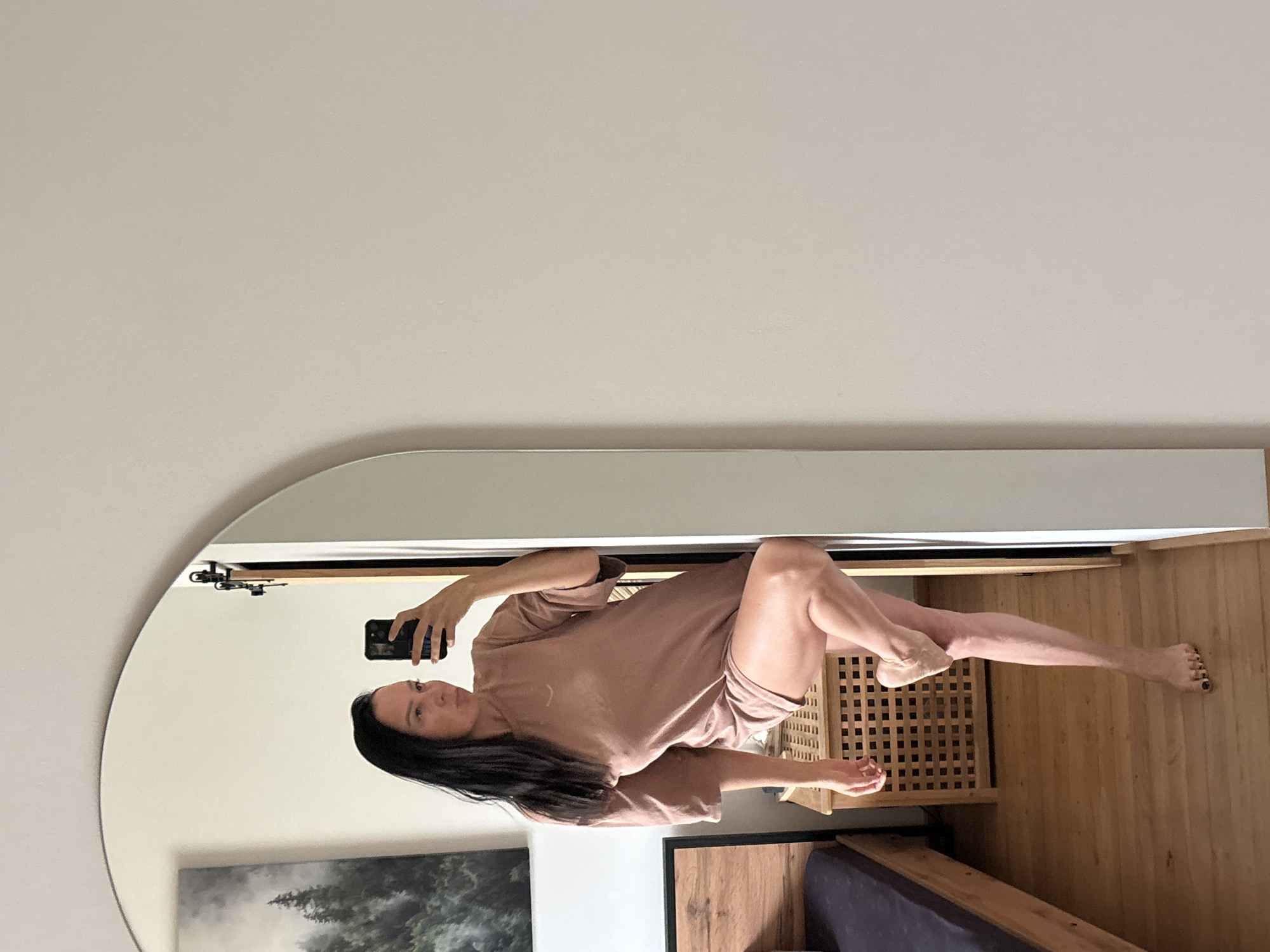 A woman is taking a selfie in a mirror in what appears to be a bedroom. She is wearing a loose, earth-toned t-shirt dress and is standing with one leg bent and resting on a wooden cabinet. The mirror is large and arched, and it reflects the woman, a portion of a wooden bed frame, and a painting of a forest scene on the wall. The room has wooden floors and light-colored walls. The image does not contain information to determine the location in Chaikovsky, Russia.