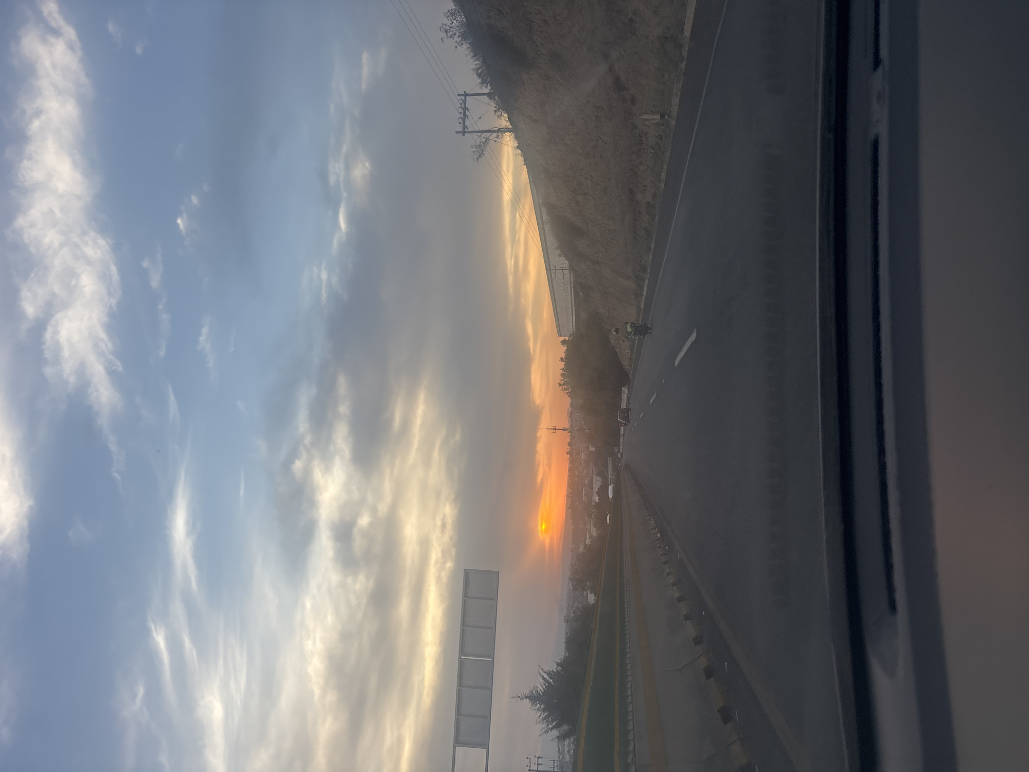 The image depicts a highway scene at sunset, viewed from inside a moving vehicle. The sky occupies a significant portion of the frame, showcasing a gradient from light blue-grey with scattered wispy white clouds towards the upper left, transitioning to vibrant orange and yellow hues near the horizon on the right. The sun, a prominent orange orb, is low on the horizon, partially obscured by haze or clouds, illuminating the sky with a warm glow.
To the right, a multi-lane asphalt highway extends into the distance, marked with distinct white and yellow lines. Alongside the highway, a steep, dry hill with sparse vegetation rises, featuring utility poles and power lines running along its slope. Further down the road, several vehicles appear as dark silhouettes against the bright sunset, and a person wearing a high-visibility vest is visible near the road's shoulder. A large, blank rectangular sign structure is silhouetted against the colorful sky in the mid-ground. The right edge of the image shows a partial view of a vehicle's interior frame. The scene captures the tranquil moment of dusk along a road, consistent with a setting in an area like Cuautitlán Izcalli, Mexico. No discernible text is visible.