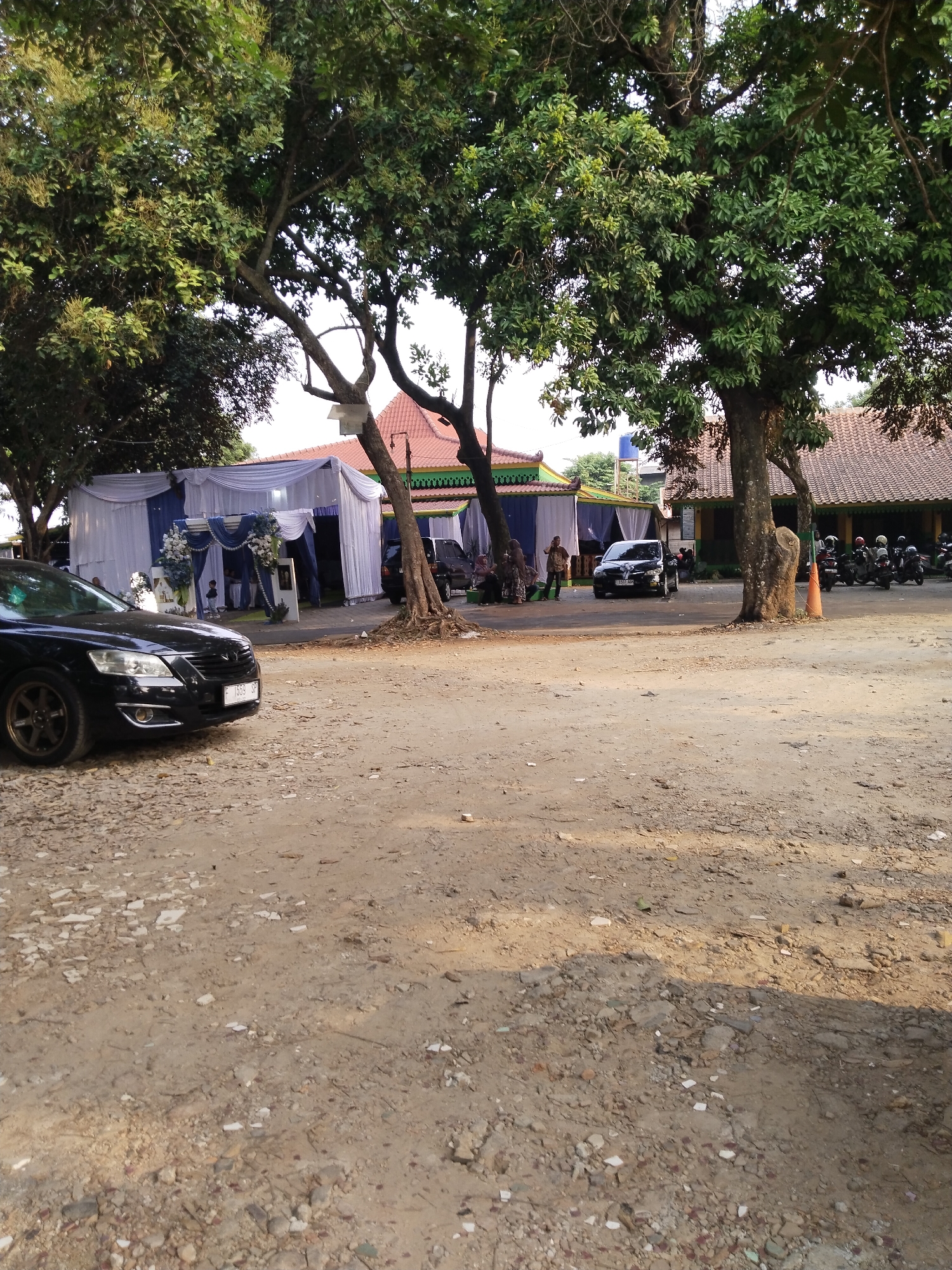 The image shows an outdoor scene, likely a gathering or event. A white tent with blue accents is set up, possibly for a wedding or reception. Several people are visible, some standing near the tent and others seated. There are at least two cars in the foreground and background, and a row of motorcycles is parked on the right side of the image. The ground is unpaved, with dirt and gravel, and there are large trees providing shade. The architecture of the buildings in the background suggests a residential or community setting.