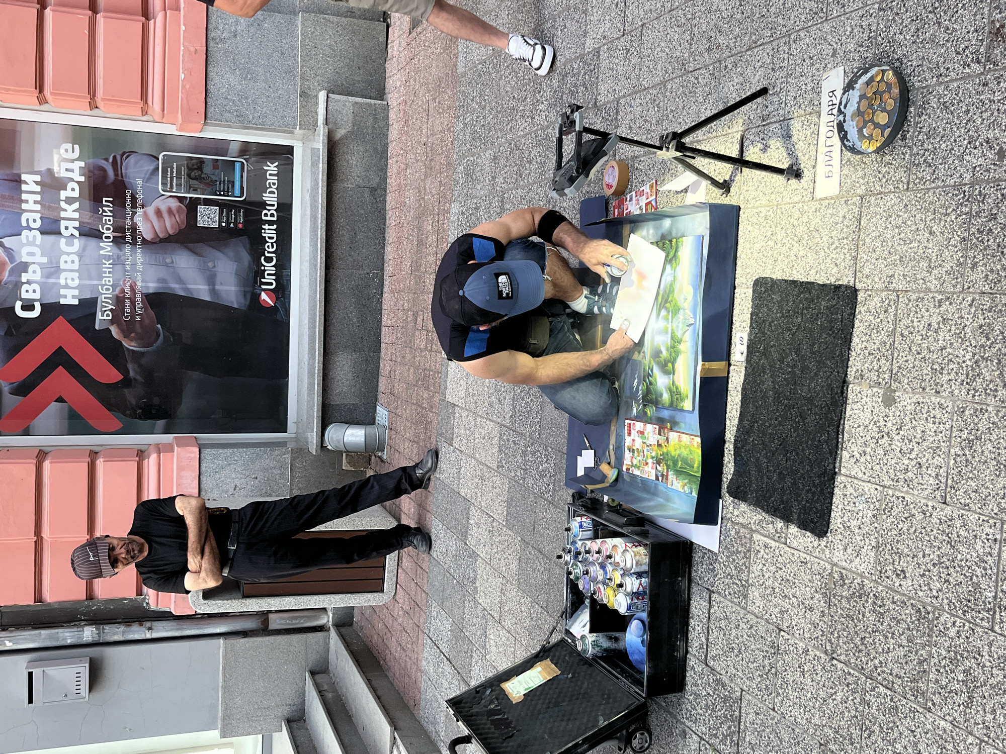 An outdoor street scene in Plovdiv, Bulgaria, features an artist spray-painting a mural on the sidewalk. The artist, wearing a black t-shirt, blue jeans, and a baseball cap, is crouched down, using spray cans from an open black case. The mural depicts a landscape scene with trees and water. A tripod is set up next to the artist, possibly for a camera or lighting. In the foreground, a collection of coins is displayed in a dish, with a sign that reads "БЛАГОДАРЯ" (Thank you) and "€ 10." To the left, a man in a black t-shirt and pants stands with his arms crossed, wearing a striped baseball cap. Behind the artist, a large advertisement for UniCredit Bulbank is displayed on the side of a building. The sidewalk is paved with granite tiles, and a dark mat is placed in front of the artist's work area. The overall atmosphere suggests a public space in a city, with artistic activity taking place.