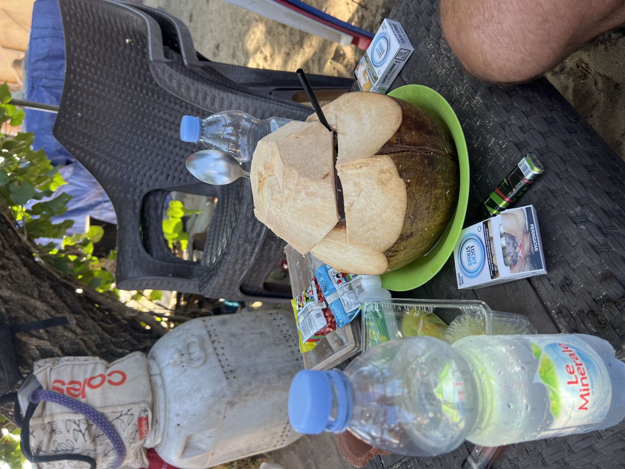 On a dark brown woven plastic table in Kuta, Indonesia, a partially opened coconut sits on a green plate. A black straw extends from the coconut. To the left of the coconut is a bottle of Le Minerale bottled water, condensation visible on its surface, and a clear plastic container holding what appears to be limes. To the right of the coconut, a pack of Lucky Strike cigarettes, open, reveals its contents, and next to it is a vape pen. In the background, a dark brown plastic chair is visible, as well as a large white container with the "Coles" logo on a beige tote bag. A tree with green leaves is overhead, casting shadows. The ground appears to be sandy.