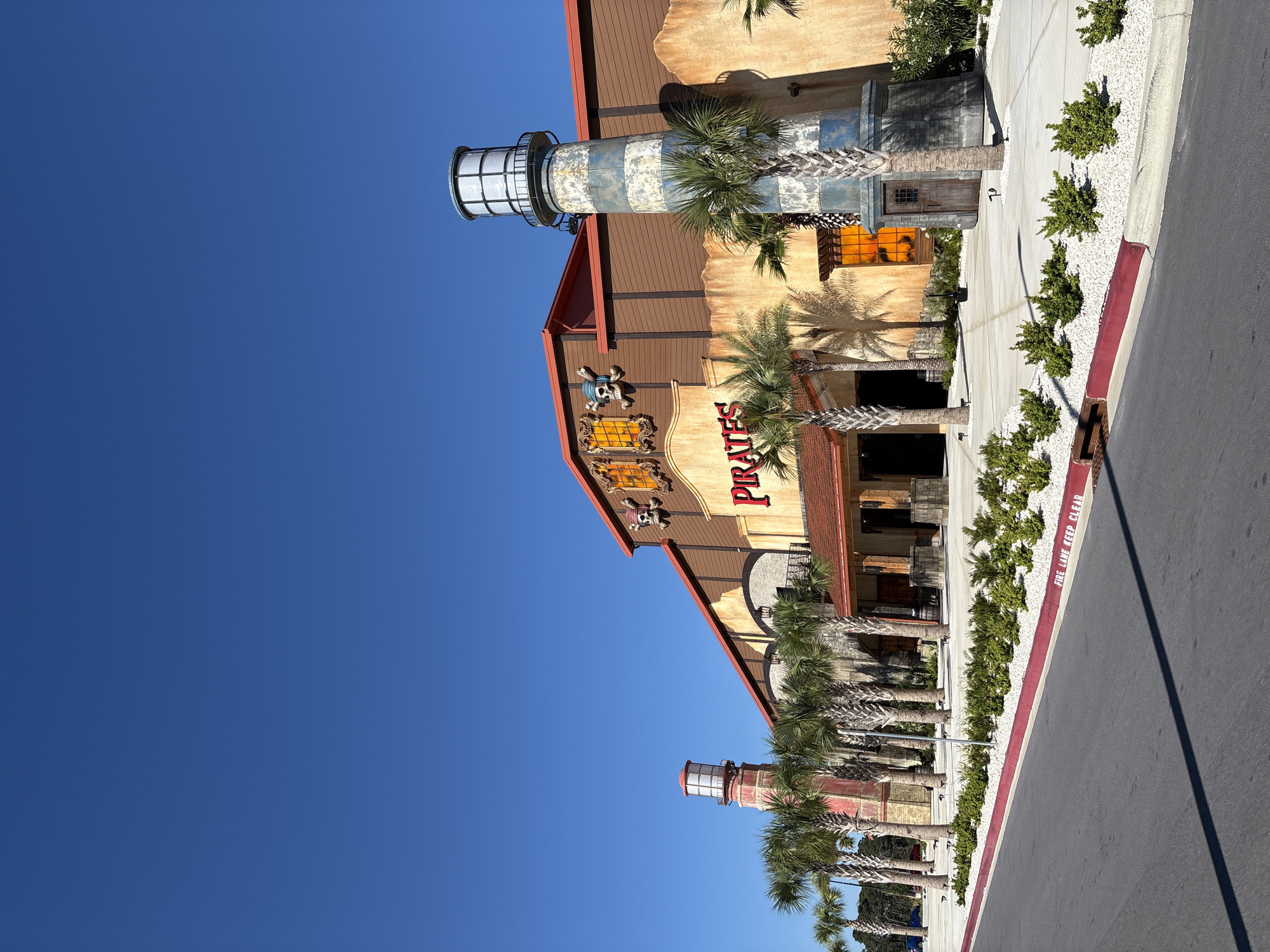 The image shows the exterior of a building called "Pirates" in Panama City Beach, Florida, USA. The building has a themed design with a lighthouse structure on each end, palm trees in front of it, and a sign that reads "Pirates". The sky is a clear blue, and the road in front of the building is visible with a designated fire lane. There is no visible presence of people.