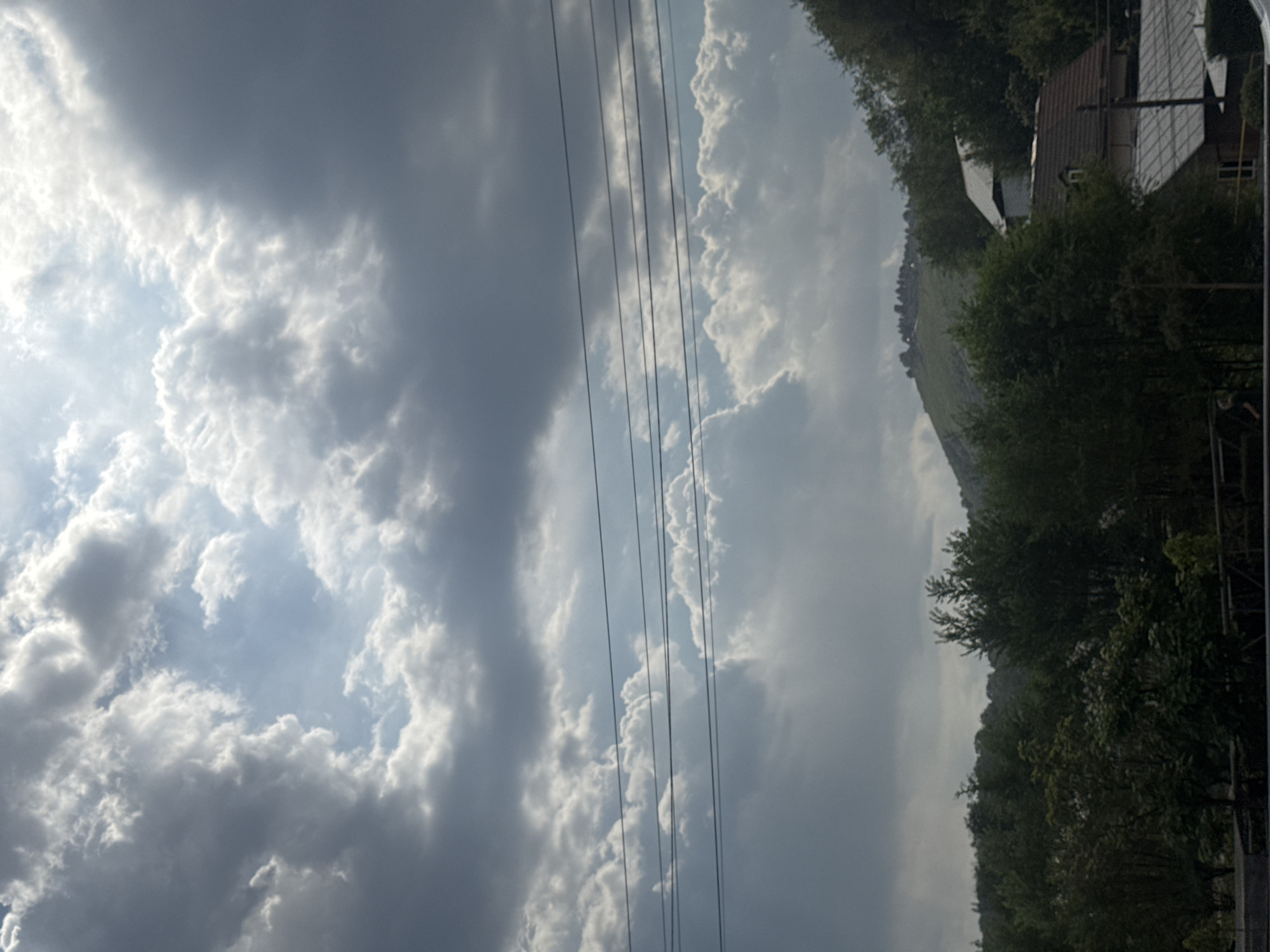 The image shows a view of a cloudy sky with the sun breaking through. In the foreground, there are several power lines stretched across the frame. Below the power lines, there is a dense line of trees and foliage, with some houses partially visible amongst them. In the background, a green, tree-covered hill rises, with a few small structures or buildings visible on its summit. The overall impression is of a semi-rural or suburban area, possibly on the outskirts of a city, with a dramatic sky. The image does not provide specific details about the city of Almaty, Kazakhstan.