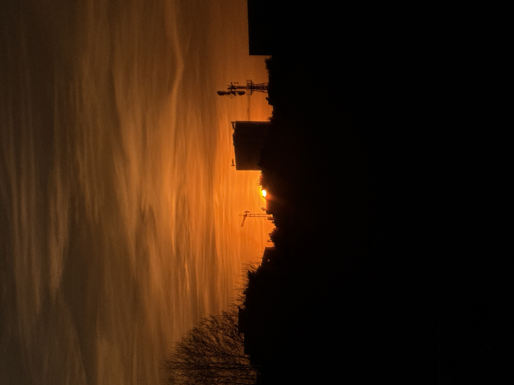 The image depicts an urban skyline in Vila Nova de Gaia, Portugal, silhouetted against a vibrant sunset or sunrise. The sky dominates the upper two-thirds, displaying a gradient from dark reddish-brown at the top to intense orange and yellow hues near the horizon, streaked with horizontal clouds. The sun, a brilliant orange-yellow disk, is partially visible just above the silhouetted horizon line, slightly left of center. The lower third of the image consists of a dark, almost black silhouette of the cityscape. Prominent features include a large, bare deciduous tree on the far left. Towards the center-left, a construction crane is visible. In the center-right, a large, multi-story building with an angled roofline stands next to a distinct communication tower equipped with multiple antennae and dishes. Another large, rectangular building is partially visible on the far right. No people or discernible actions are depicted.