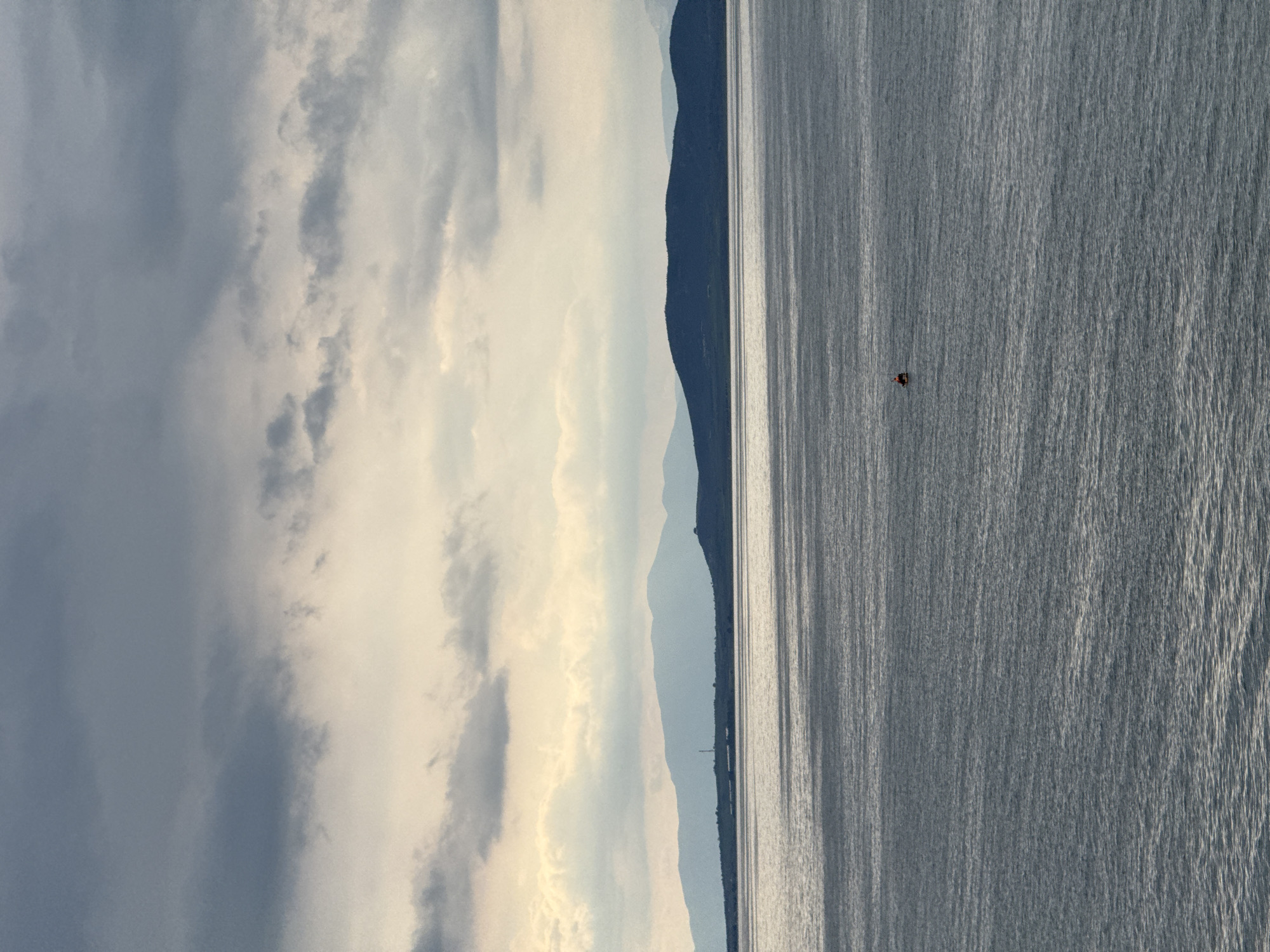 The image depicts a body of water, likely a sea or large lake, with a mountainous landmass visible along the horizon line. The sky is filled with a mixture of dark and light clouds. A small, dark object appears in the water, possibly a boat or person. The location is Fanari, Greece.