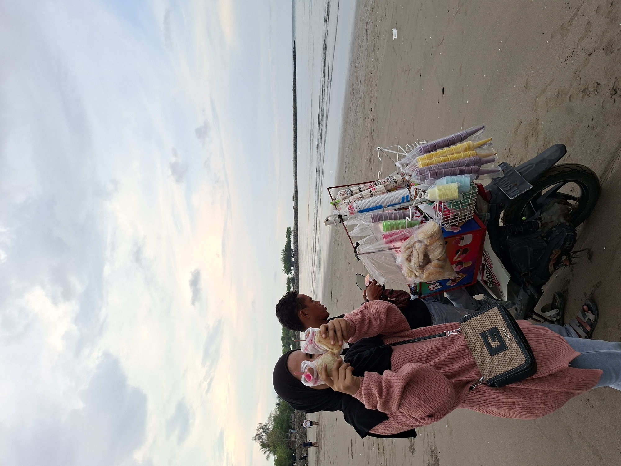 A woman wearing a pink sweater and a black hijab and a man also wearing a pink sweater stand on a sandy beach, both holding and eating what appears to be sandwiches. Next to them is a motorcycle loaded with containers of ice cream and related supplies, likely for sale. The beach stretches out to the ocean under a partly cloudy sky. The location is Lhokseumawe, Indonesia.