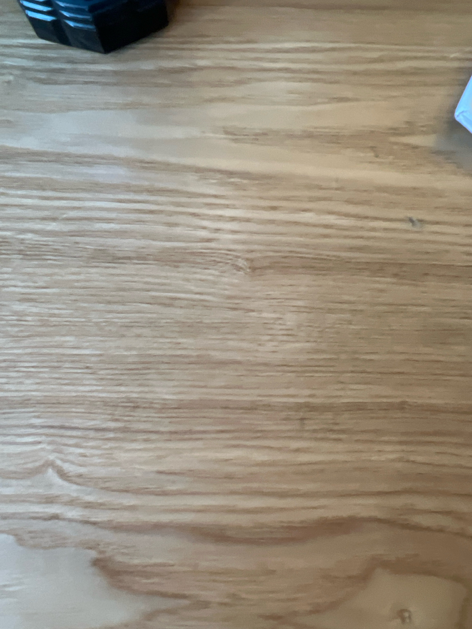 The image shows a close-up view of a light-colored wooden surface with prominent grain patterns. The wood appears to be a table or desk, with some lighter and darker streaks creating a natural, textured look. In the upper left corner, a dark, rectangular object with blue accents is partially visible. The top surface of this object shows a grid-like pattern or segmented design. In the upper right corner, a small portion of a white object with a light blue border is visible. The majority of the image is occupied by the wooden surface, which has a smooth finish. There is no indication of people, actions, or interactions. The image does not provide any information about the location in Krasnoyarsk, Russia.