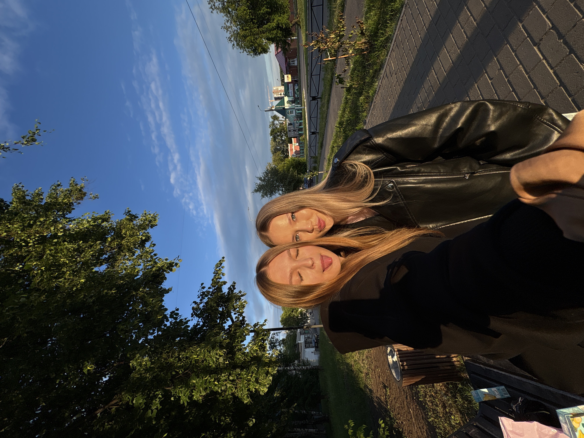 The image depicts two young women taking a selfie outdoors in Chaikovsky, Russia, during what appears to be late afternoon or early morning due to the low-angle sunlight casting long shadows.
The woman on the left, with long, light brown hair, wears a black hoodie or jacket. Her eyes are closed, and her tongue is slightly extended in a playful gesture. The woman on the right, with long blonde hair and blue eyes, wears a black leather jacket and looks towards the camera with a slight smile or pout. Her right hand, holding the camera, is visible in the lower right foreground. Both women's faces are illuminated by direct sunlight.
The immediate foreground features a paved sidewalk of rectangular blocks and a wooden slatted bench, partially obscured on the left, with a pink bag and a patterned box resting on it. A wooden trash receptacle stands next to the bench. Dense green trees and foliage occupy the left side of the image and extend into the mid-ground, forming a park-like environment alongside the sidewalk.
Beyond a dark metal fence and a street, various buildings line the background. A prominent light-colored building with a distinctive green roof features Cyrillic text "МАГАЗИН" (Shop) and "МОЛОКО" (Milk). To its right stands a reddish-brown building with white trim. Further in the distance, a construction site with a visible crane is present. A white single-story structure is partially hidden by trees to the left of the green-roofed building. A street lamp post is visible between the road and the buildings. The sky is clear blue with scattered white, wispy clouds, and power lines are visible in the upper right quadrant.