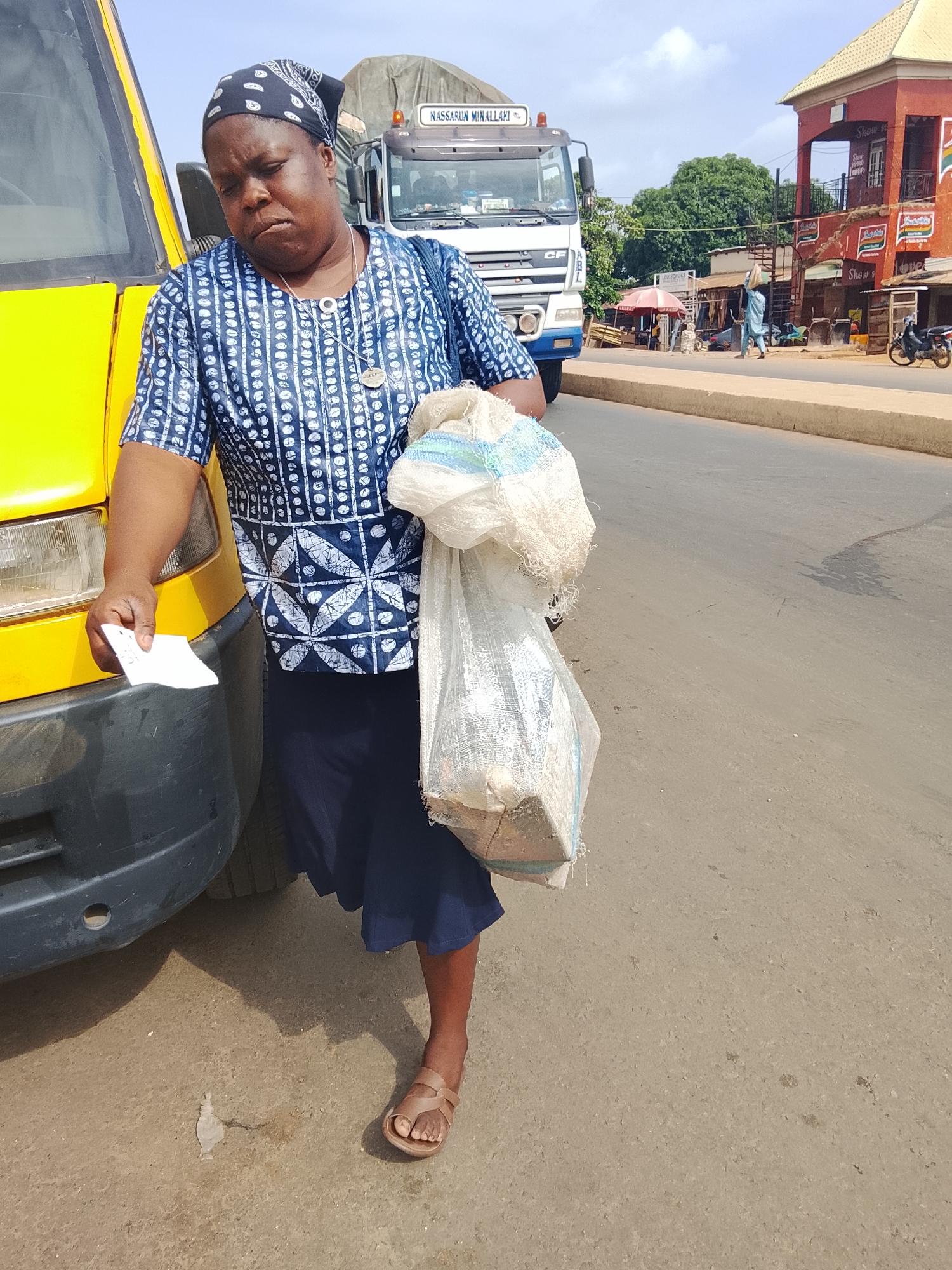 An outdoor, eye-level, medium shot captures a woman standing by a yellow bus on a street in Akwanga, Nigeria. The woman, wearing a blue and white patterned tunic, dark blue skirt, and a black headscarf, holds a white plastic bag in her right hand and a crumpled white piece of paper in her left. Her head is bowed, and her expression is somber. Behind her, a white truck with the words "NASSARUN MINALLAHI" is visible, along with buildings and trees. The street is paved, and a beige curb runs along the right side. The time of day appears to be daytime, with clear skies and shadows indicating the sun is high.
