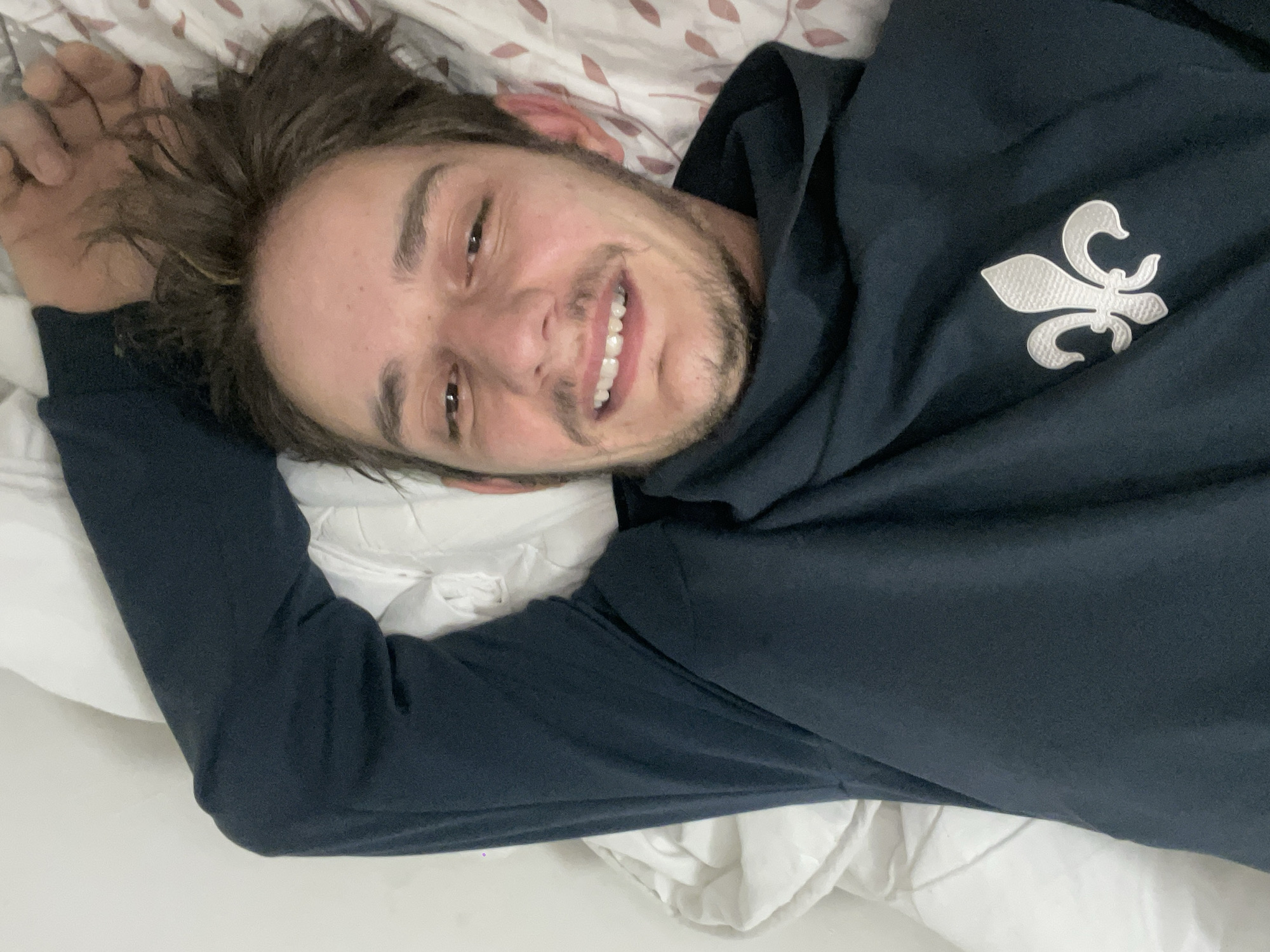 A medium shot shows a man lying on a bed, smiling directly at the camera. He has brown hair and a short beard. He is wearing a navy blue hoodie with a white fleur-de-lis design on the right side of the chest. The bed has white sheets and a patterned pillowcase. The location appears to be indoors in Unknown, Unknown.