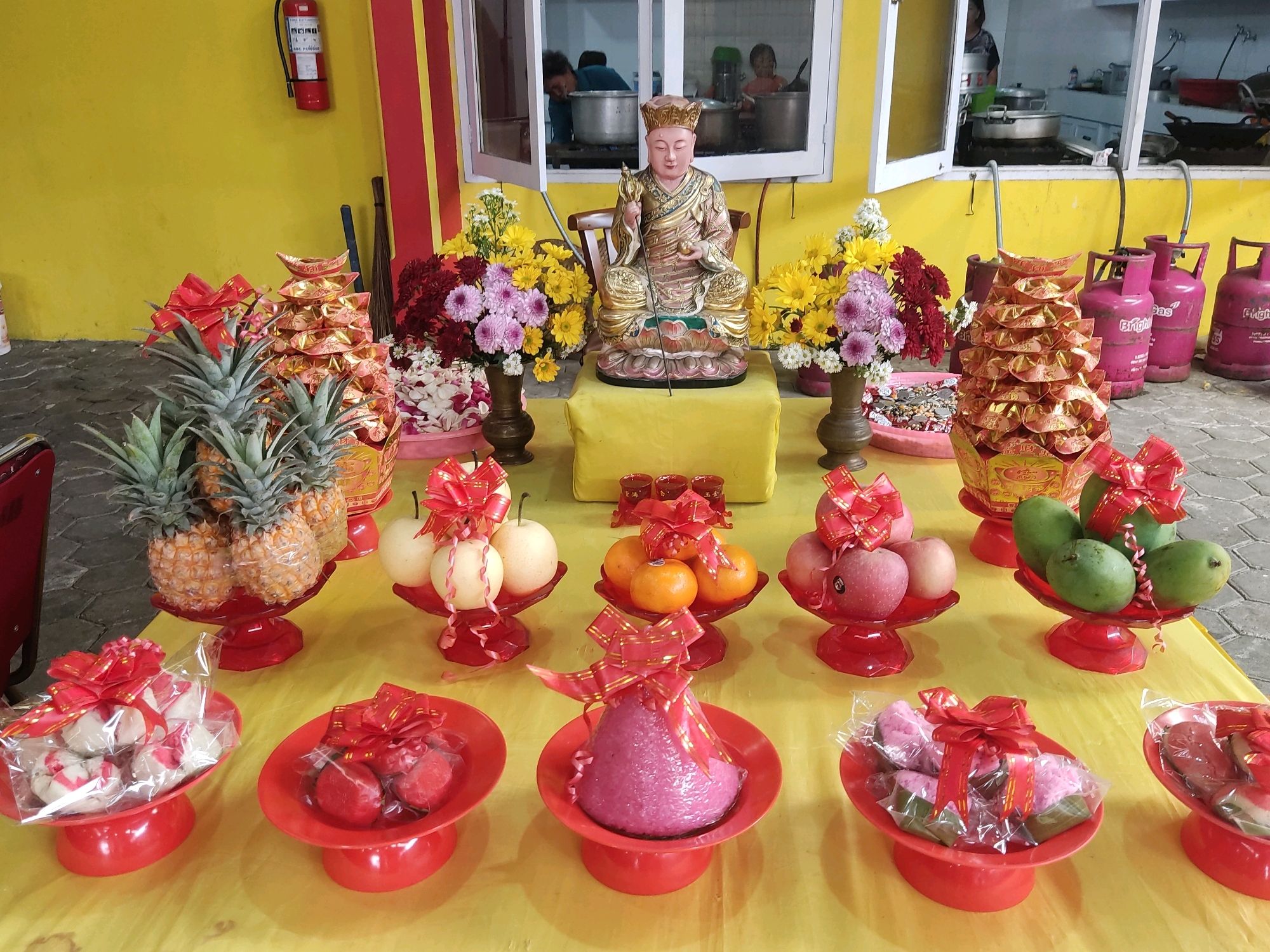The image depicts an altar laden with offerings in what appears to be an outdoor or semi-outdoor setting, possibly at a temple or a private residence, in Magelang, Indonesia. The background features a bright yellow wall with red and yellow vertical stripes, and a window revealing people in what looks like a kitchen environment, with cooking pots on stoves. To the left, a red fire extinguisher is mounted on the wall.
The altar itself is covered with a yellow cloth. Prominently displayed at the center is a seated Buddha statue adorned in golden robes and a crown, holding a scepter and a spherical object. Flanking the statue are vases filled with various colorful flowers, including red dahlias, pink chrysanthemums, and yellow daisies.
In front of the statue, and arranged symmetrically across the altar, are numerous offerings. These include stacks of gold-colored paper ingots, commonly used in offerings, several red pedestals holding an assortment of fruits: pineapples, pears, oranges, apples, and mangoes. Some of the fruits are whole, while others are sliced and presented in clear plastic packaging, each tied with red ribbon. There are also offerings of what appear to be sweet treats or cakes, some pink and white, and others red, also packaged and adorned with red ribbons. In the foreground, several more red pedestals hold these packaged treats.
The overall scene suggests a religious or ceremonial occasion, likely a form of worship or prayer involving offerings. The presence of the Buddha statue and the nature of the offerings are indicative of Buddhist traditions. The environment seems to be a functional space, possibly connected to a community kitchen, given the view through the window.