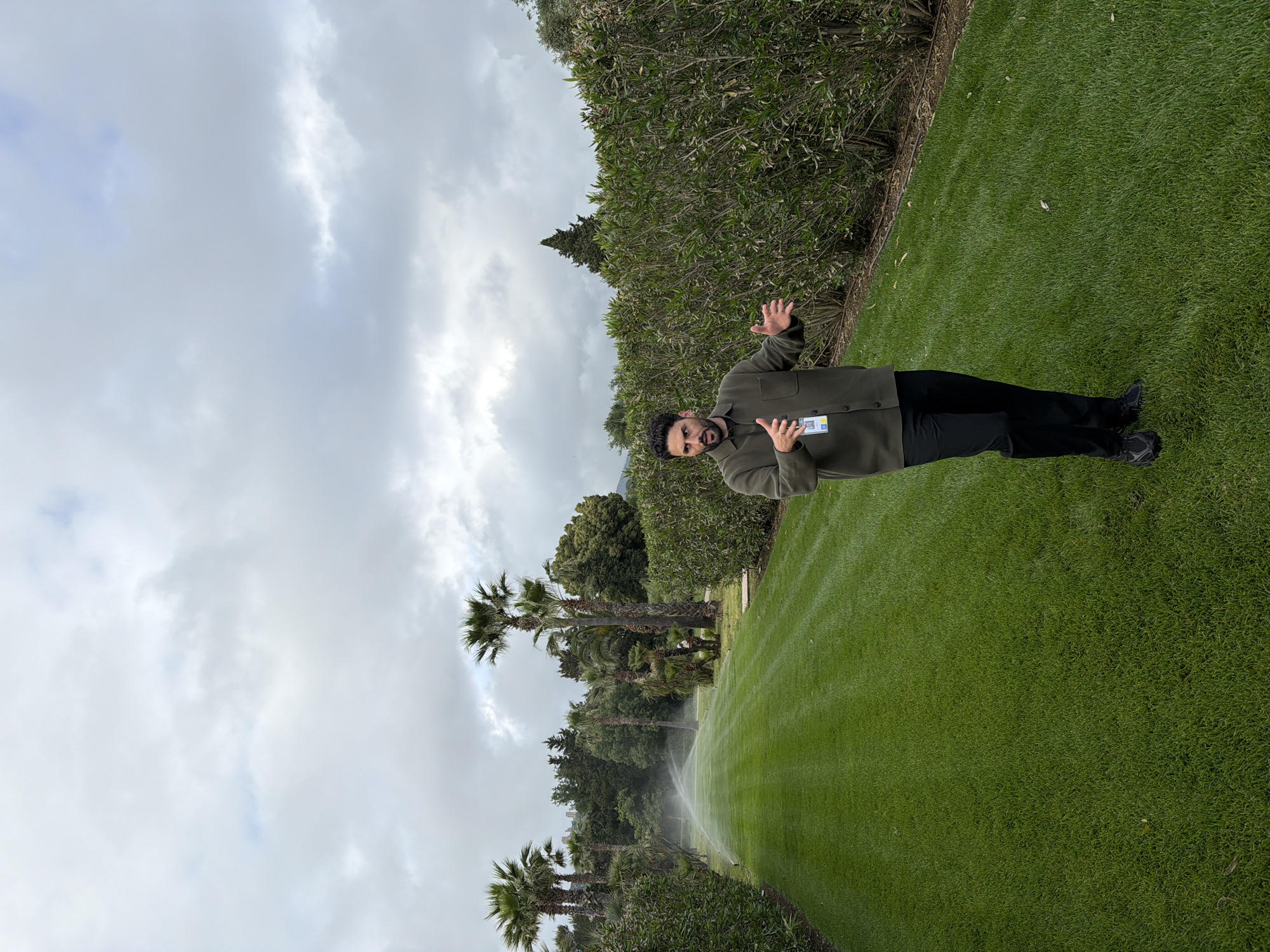 A person stands on a large green lawn in Cogolin, France. The sky is overcast. The grass is neatly trimmed, with lines from mowing visible. Palm trees and dense greenery line the edge of the lawn. Sprinklers are in operation, watering the grass. The person is wearing a dark jacket and pants, and making hand gestures.