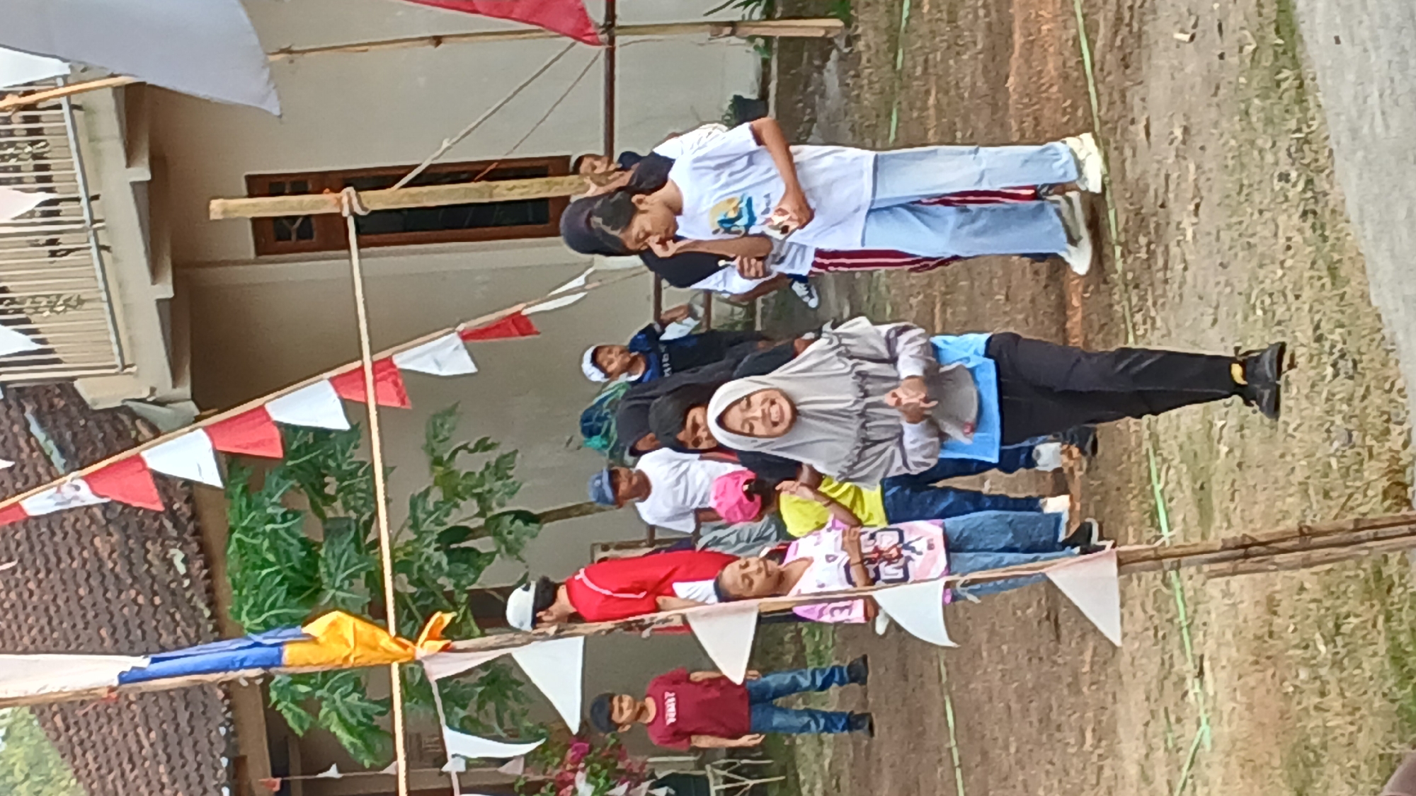 A group of children and adults are gathered outdoors in what appears to be a village setting in Mendut, Indonesia. The scene is decorated with red and white triangular flags strung across a makeshift line, possibly for a local celebration or event. In the foreground, a young girl wearing a grey hijab and blue top is the primary subject, her hands clasped in front of her and a wide smile on her face. To her left, another girl in a pink and white shirt and jeans stands looking forward. Behind them, a boy in a red t-shirt and jeans is partially visible. Further back, several other children and adults are standing and interacting. The environment consists of a dirt ground with sparse dry grass, and a building with a tiled roof in the background. A wooden pole with bamboo support stands in the middle ground, draped with fabric and flags. The overall atmosphere seems to be festive and casual.