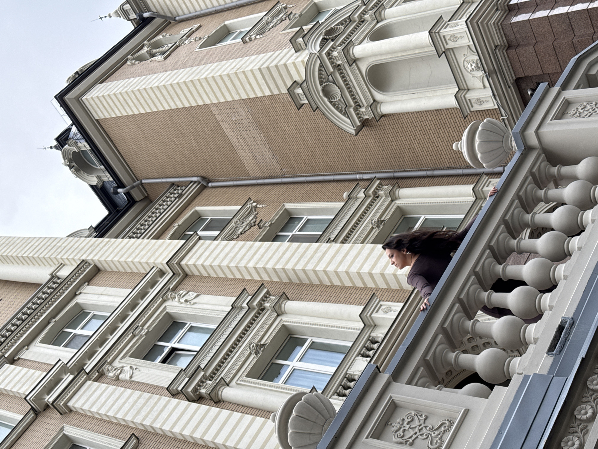 A low-angle, full-shot photograph captures a woman leaning on a decorative balcony railing in front of an ornate building. The woman, with long dark hair, is looking upwards and to her left, her head tilted back. She wears a dark-colored, long-sleeved top. The building behind her features a facade of light pink brick with white vertical stripes and decorative architectural elements, including columns and arched window frames. The sky above is overcast and grey. The image appears to be taken in Kazan, Russia, given the architectural style of the building which is reminiscent of historic Kazan structures.
