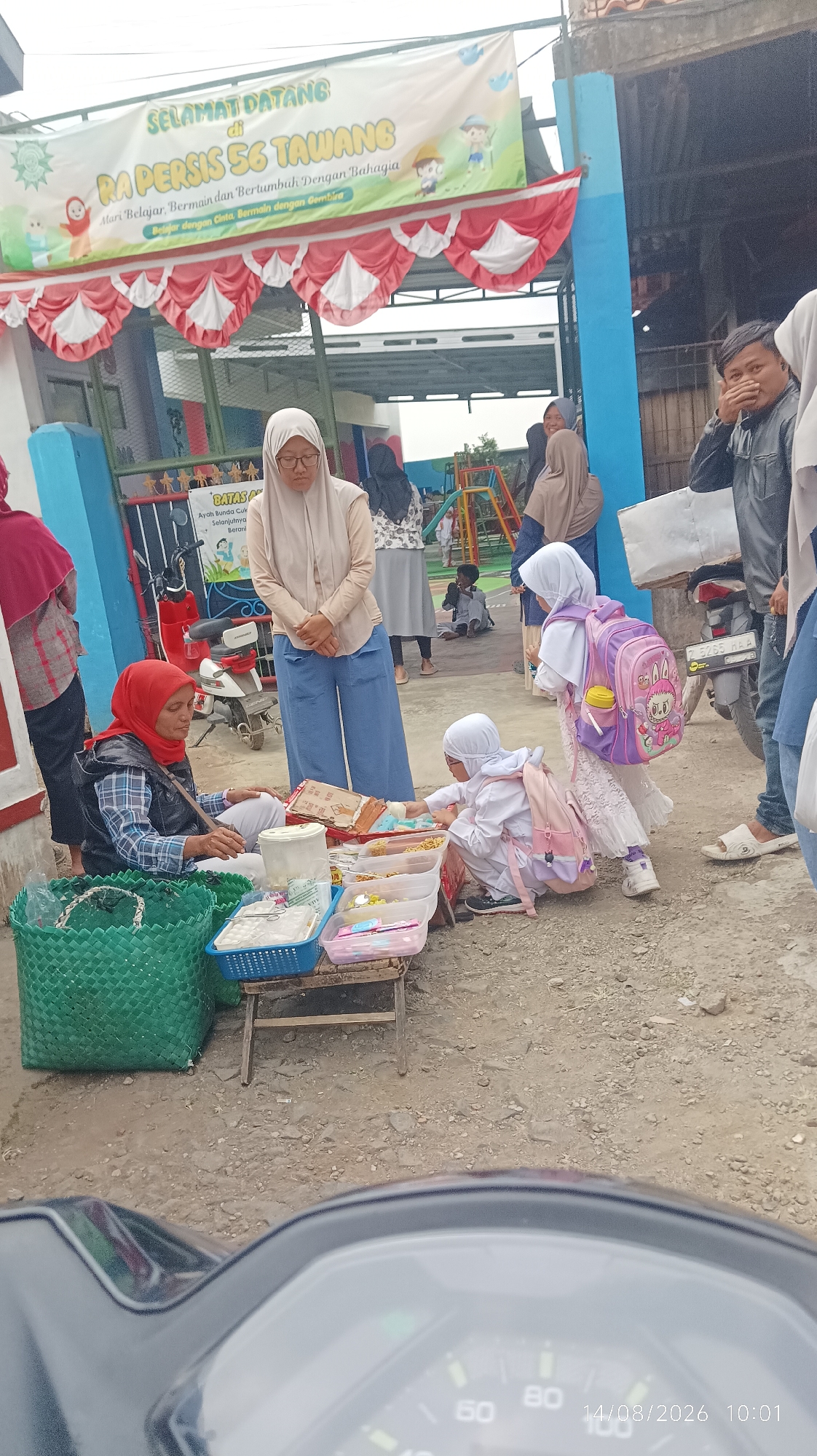 The image depicts an outdoor scene likely in Tasikmalaya, Indonesia, based on the provided context. In the foreground, a person is seated next to a small cart laden with various items, possibly snacks or small goods, displayed in plastic containers. Two young children, dressed in white, are interacting with the items on the cart. One child is reaching for an item, while the other, wearing a pink backpack, stands behind. A woman in a red headscarf is seated on the ground near the cart, appearing to be associated with the vendor.
In the mid-ground, several adults and children are present. A woman in a beige hijab and blue pants stands near a red and white striped awning. Further back, more people are visible, some walking and others gathered. Behind them, a colorful playground structure with slides can be seen. A motorcycle is parked to the right of the frame, with a man in a dark jacket standing nearby, his hand near his face.
The backdrop features a building with blue and white paint. A large banner with Indonesian text, including "SELAMAT DATANG di RA PERSIS 56 TAWANG" (Welcome to RA Persis 56 Tawang), is prominently displayed above the awning. The ground is covered with gravel and dirt. The overall scene appears to be a public gathering or an event outside an educational institution. The time stamp on the bottom right corner indicates "14/08/2026 10:01".