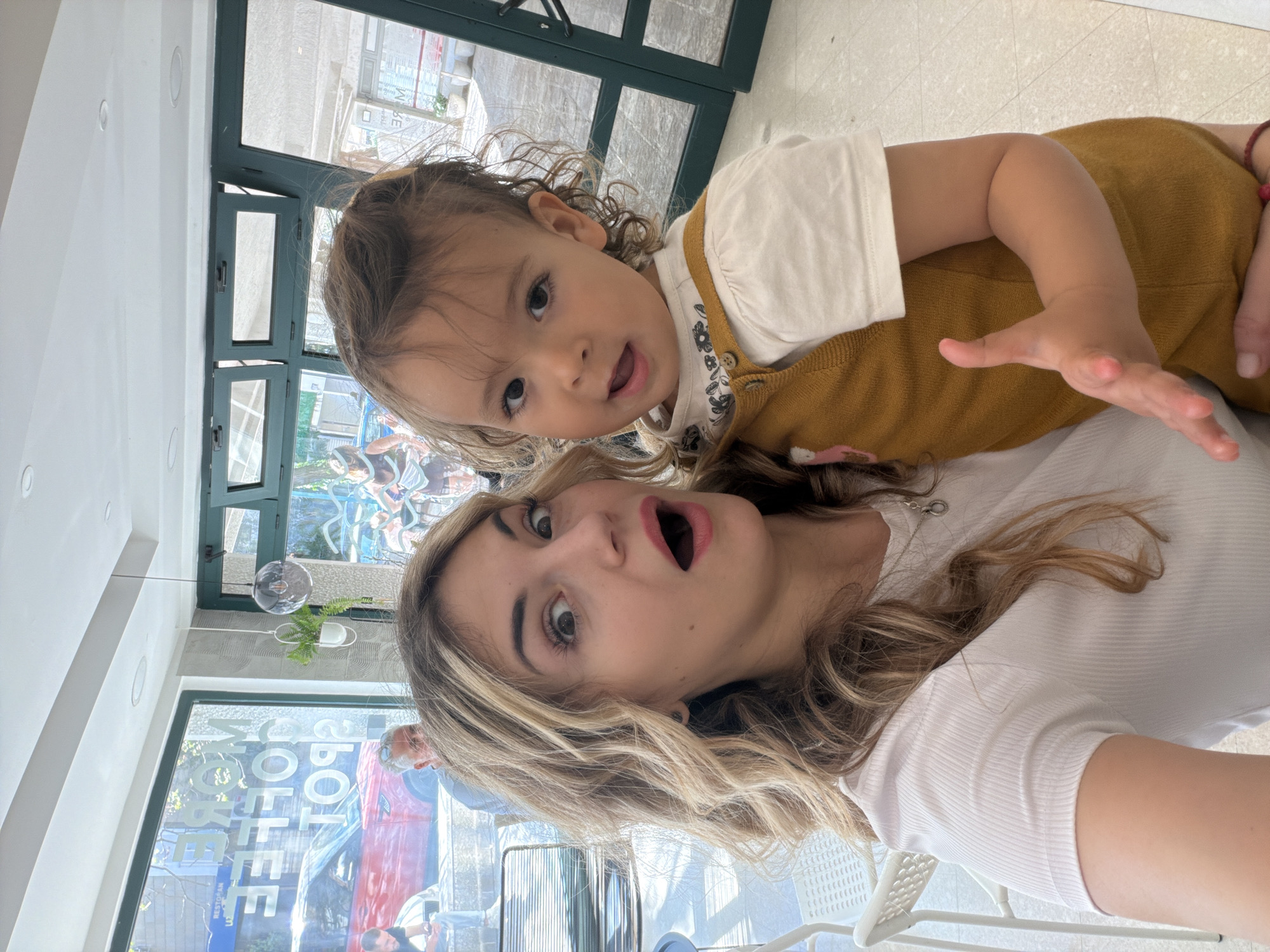 A light-skinned woman and a toddler are in a possible cafe in Budva, Montenegro. The woman has blonde hair and is wearing a white shirt. She has an open mouth as if posing for the camera. The child has brown hair and is wearing a white shirt with a brown jumper. There is outdoor seating visible through the cafe windows, along with street details and a building with the word "COFFEE" in large letters. Additionally, outdoor greenery and dark green window frames are seen.