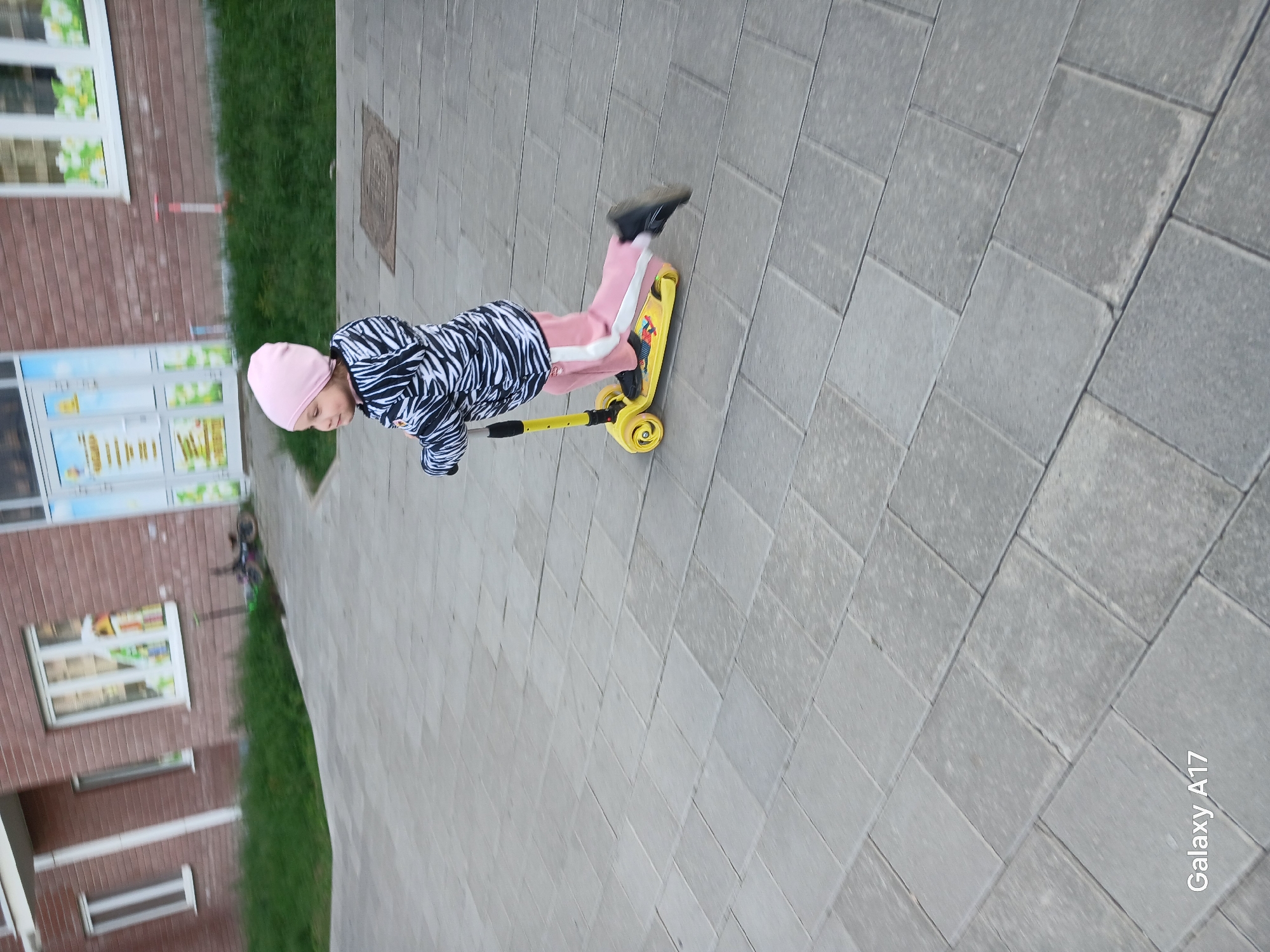 A child in a pink hat, zebra-striped jacket, and pink pants rides a yellow scooter on a gray brick-paved sidewalk. A building with brick walls, white-framed windows, and a glass door is visible to the left. A strip of green grass separates the sidewalk from the building. The location is likely in the city of Yekaterinburg, Russia.