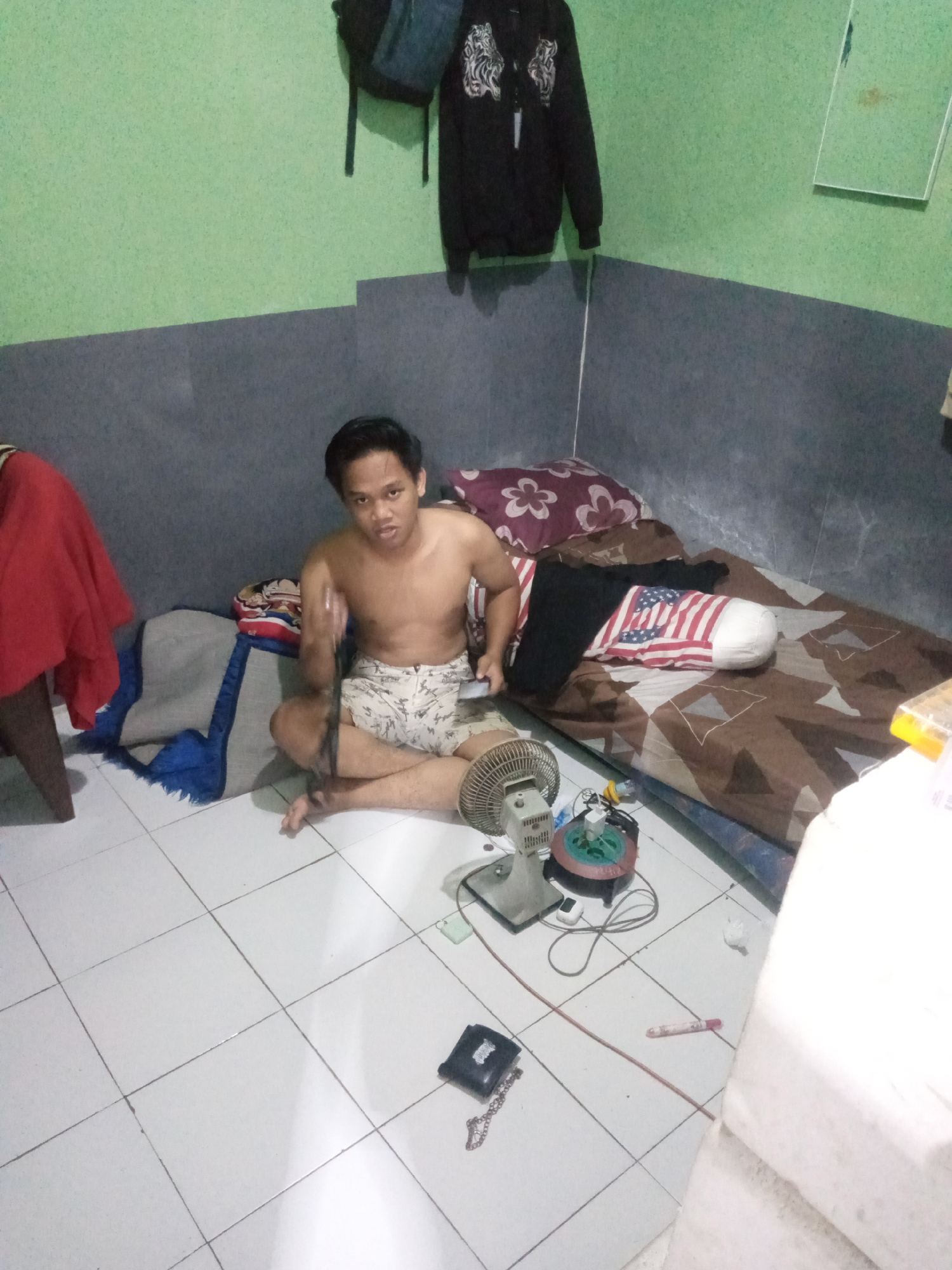 A young man with short black hair and a light complexion is sitting on a tiled floor in what appears to be a bedroom. He is shirtless and wearing patterned shorts. His legs are crossed, and his right hand is raised in front of him, holding a dark object. To his left, a red cloth is draped over a chair. Behind him, a mattress is covered with a patterned brown and white sheet, and several pillows are visible, one of which is decorated with the American flag. A small electric fan and other electronic devices are on the floor near the man. The walls of the room are painted in two different colors: a light green on the upper portion and a dark grey on the lower portion. A black jacket with a graphic design is hanging on the wall behind the man, and a backpack is visible above it. A mirror is also mounted on the wall. The floor is tiled with white squares. The overall impression is of a casual, lived-in space. This scene is located in Wana Kerta, Indonesia.