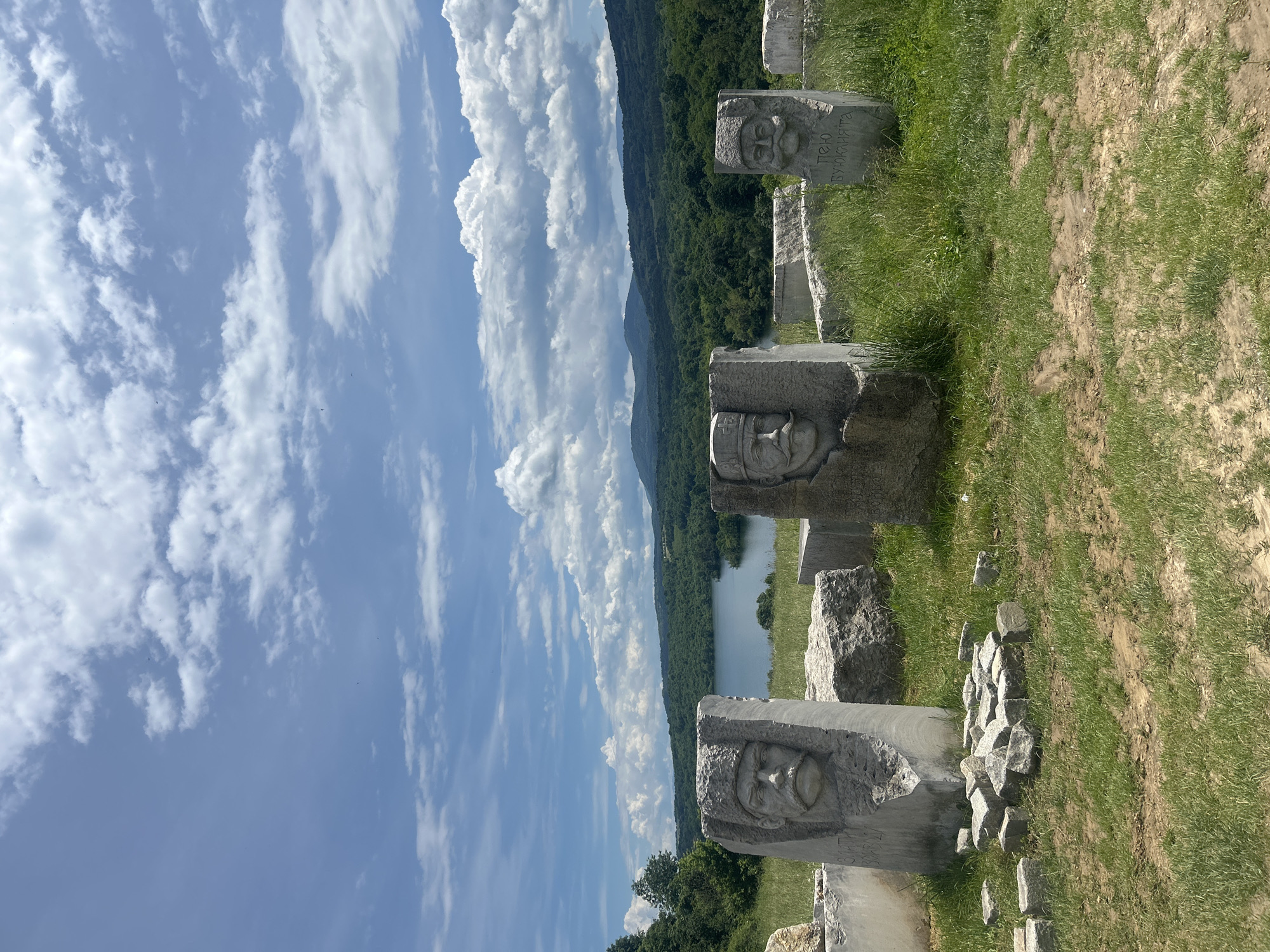 This image captures a serene outdoor scene, likely in Rayuvtsi, Bulgaria, under a bright, partly cloudy sky. The main subjects are several weathered stone sculptures arranged in a row on a grassy slope in the foreground and mid-ground.
Each sculpture is a relief carving or bust depicting a male face, some with prominent beards and traditional headwear. For instance, one sculpture clearly shows a man with a mustache and what appears to be a cap or hat. Another, further down the row, features a man with a full beard and a more ornate crown-like cap. Around the base of some sculptures, smaller, broken pieces of stone are scattered, suggesting either ongoing work or natural erosion.
Visible on one of the sculptures is Cyrillic text: "ПЕНЧО" (Pencho) and beneath it, "БУЮКЛИЯТА" (Buyukliyta), likely identifying the individual depicted or the sculptor.
Behind the sculptures, a tranquil body of water, resembling a lake or reservoir, stretches across the mid-ground. It is bordered by lush, verdant hills and mountains densely covered with trees, extending into the distant horizon. The sky is a vibrant blue, adorned with large, fluffy white cumulus clouds, indicating fair and sunny weather, likely during the day. The foreground consists of natural ground with patches of green grass and some bare earth. The scene conveys a sense of historical reflection or artistic appreciation within a beautiful natural landscape.