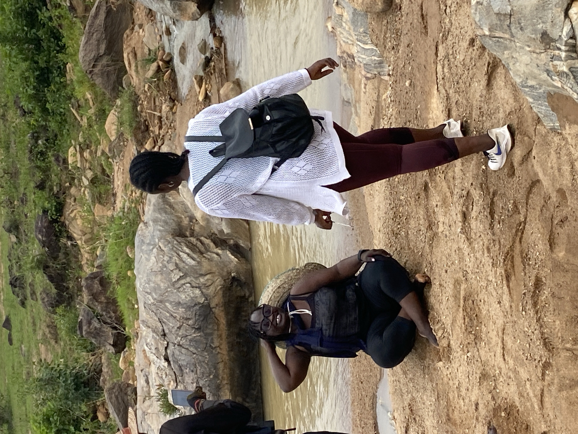 The image shows two women posing near a stream, possibly a popular tourist spot in the vicinity of Ganawuri, Nigeria. The woman standing wears a white, loosely-knit sweater, maroon leggings, and white sneakers with blue accents. She also has a black backpack. The other woman sits near the water's edge, wearing a dark top, dark shorts, and glasses. She appears to be posing with one hand near her head. The environment consists of rocky terrain and a stream with some grassy areas in the background.